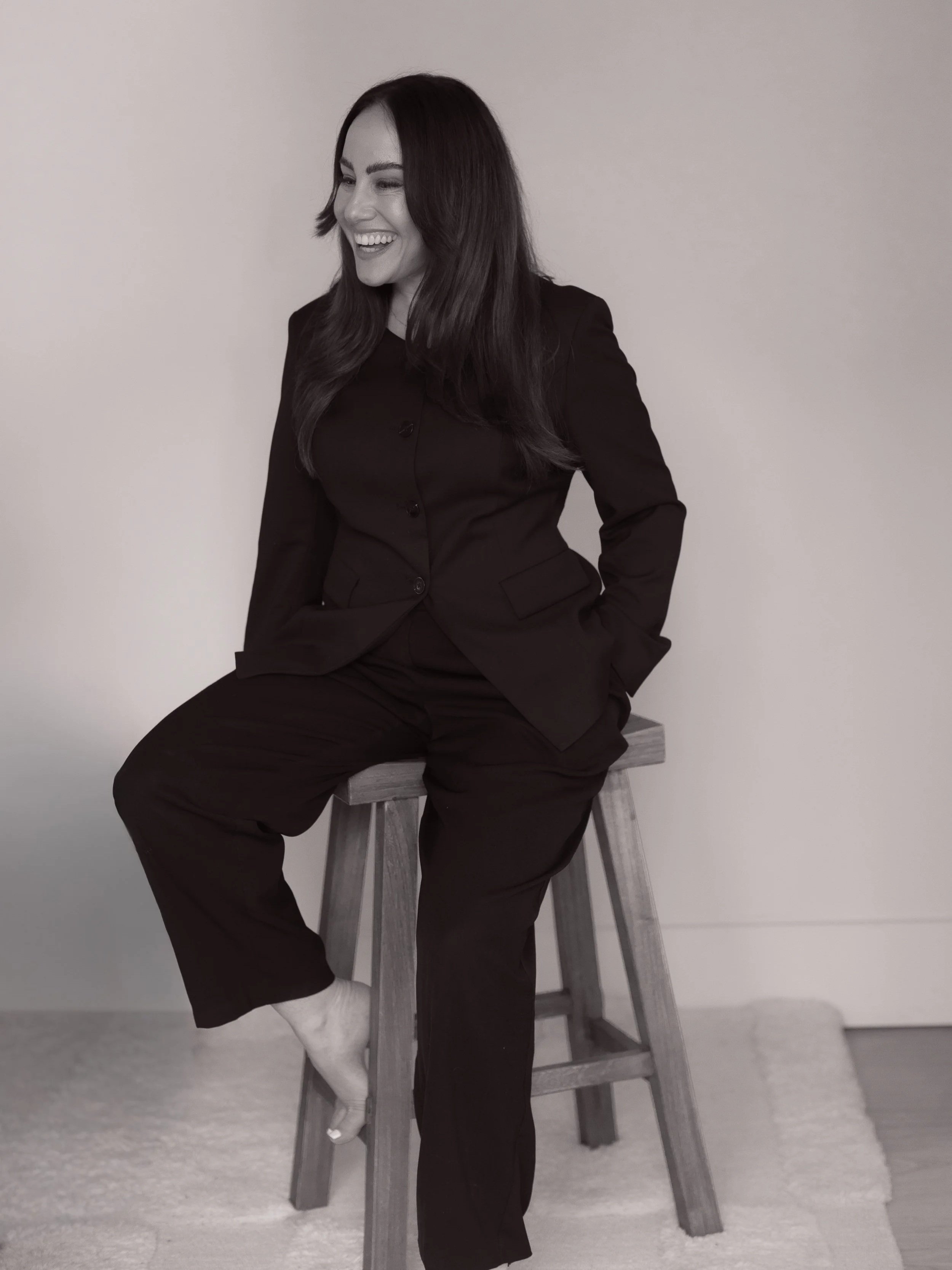 A woman with long dark hair, wearing a black suit, sitting on a wooden stool, smiling and looking to her left in a room with a light wall and a carpeted floor.
