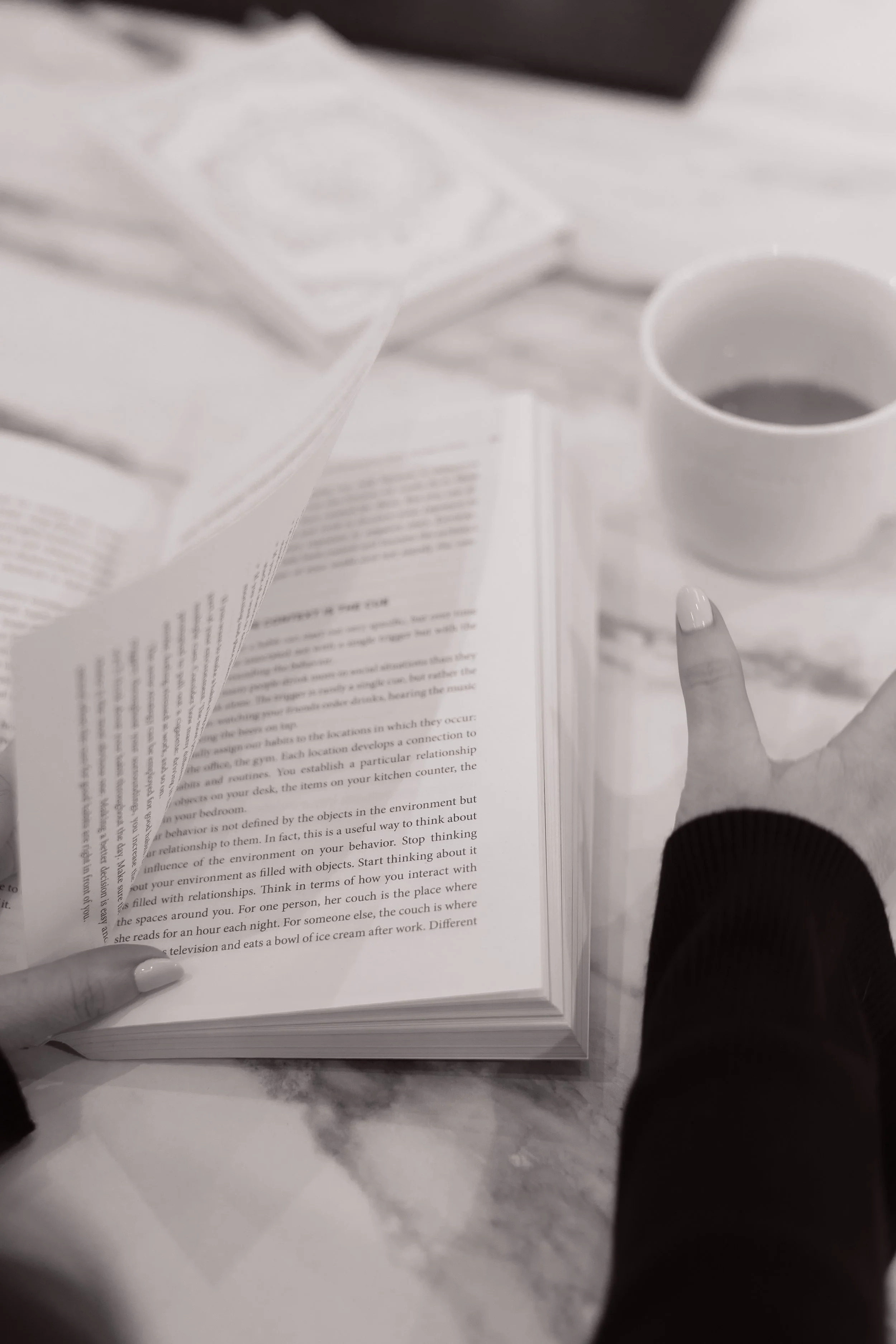 A person with painted nails is turning pages of a book on a white marble table. Next to the book is a cup of coffee and a packet of napkins.
