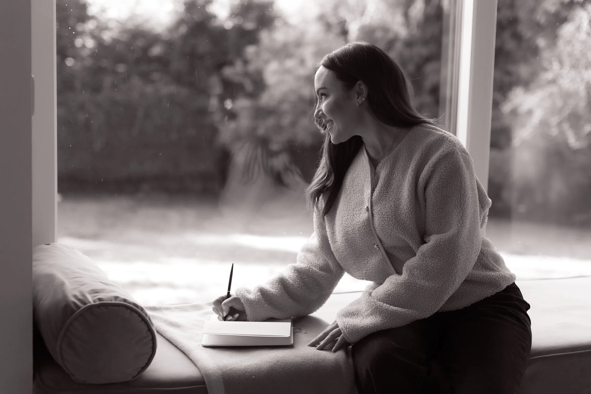 A woman sitting on a window seat with a notebook and pen, smiling and looking out the window, in a cozy setting.