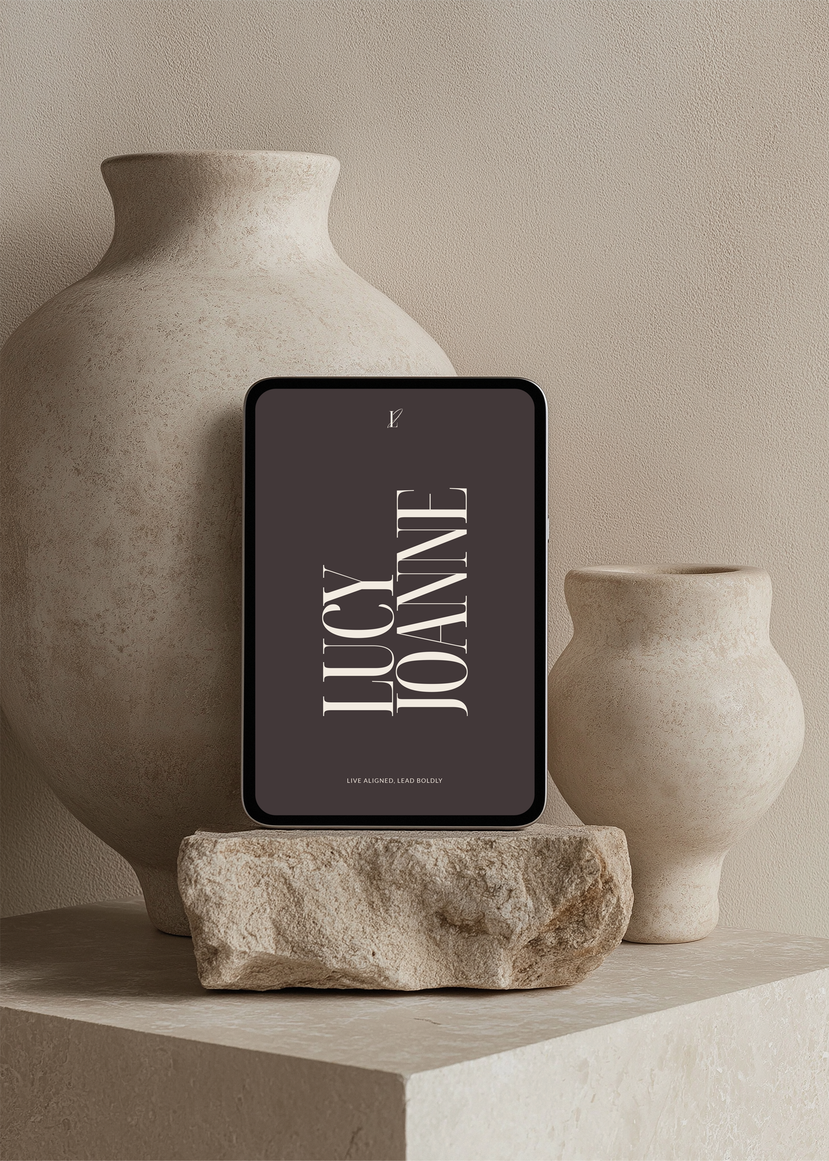 A tablet displays the text 'LUCKY JOE' and 'LIVE ALIGNED, LEAD BOLDLY' against a dark background. The tablet is placed on a rough stone and surrounded by two beige, textured vases in a neutral-colored setting.
