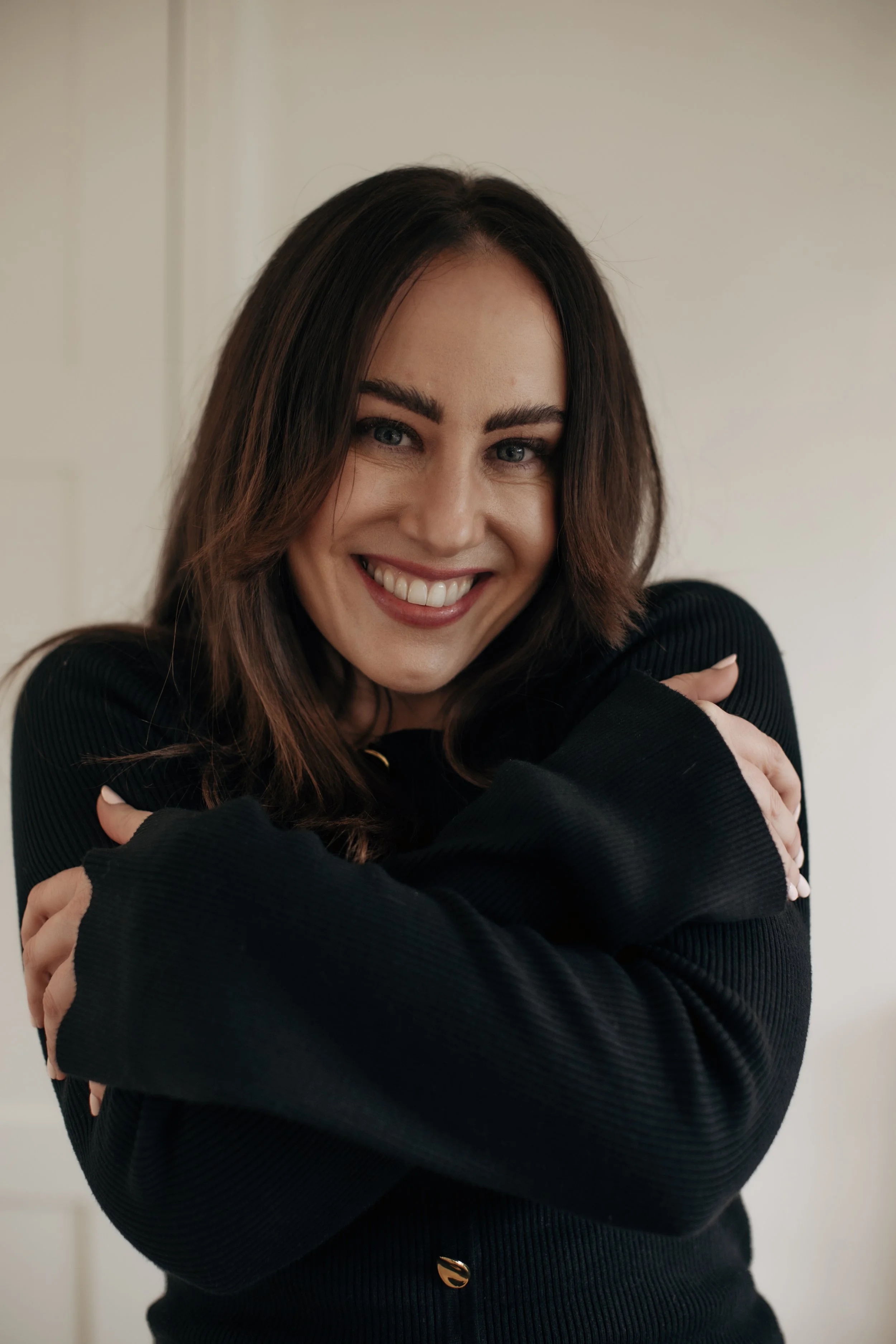A woman with shoulder-length brown hair and glowing skin is smiling and hugging herself, wearing a black sweater, against a neutral background.