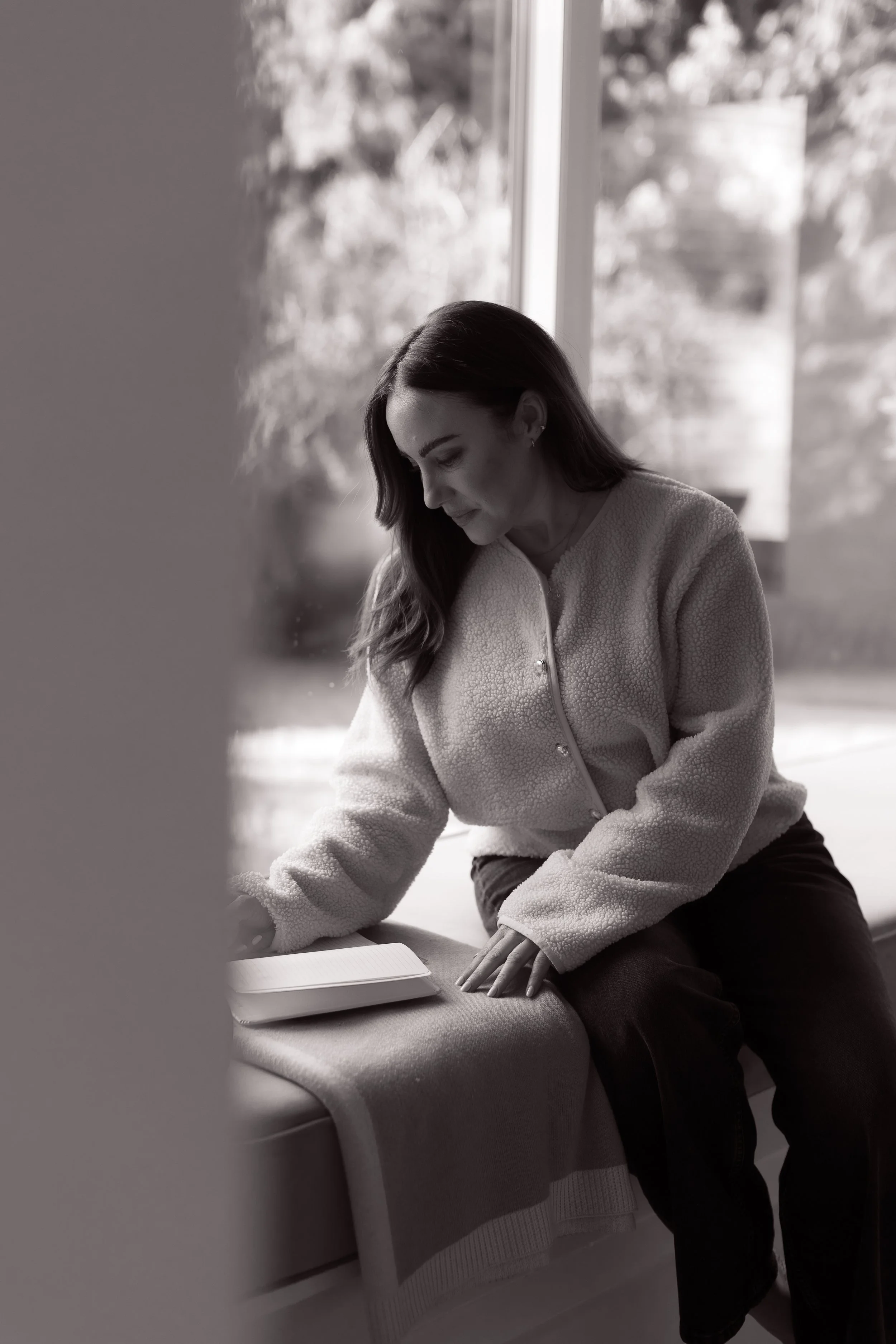 A woman with long hair, dressed in a cozy fleece jacket, sitting by a window and looking down at a book or notebook.