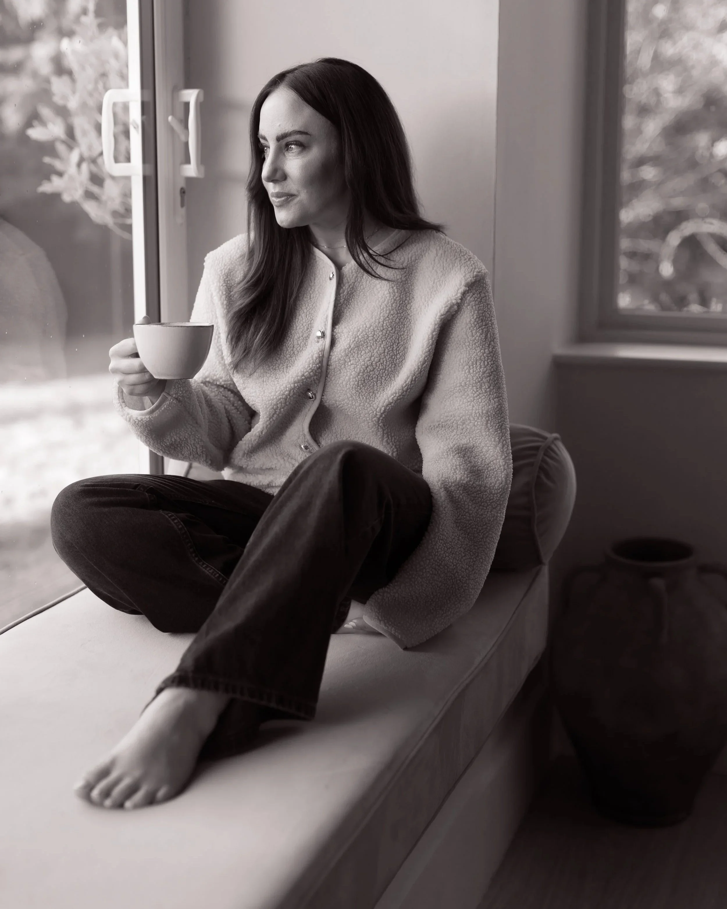 A woman sitting on a window seat, holding a cup, looking out the window, in a room with natural light.