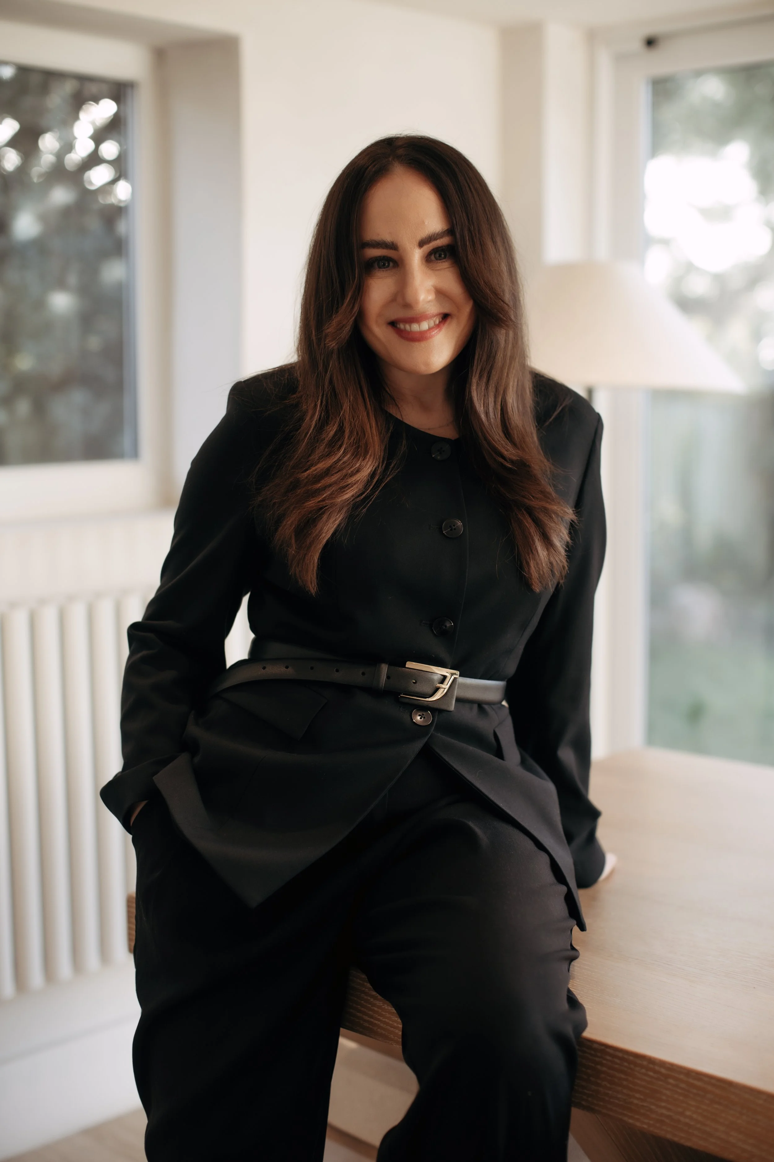 A woman with long brown hair, wearing a black suit with a belt, sitting on a wooden table in a bright room with large windows and white walls.