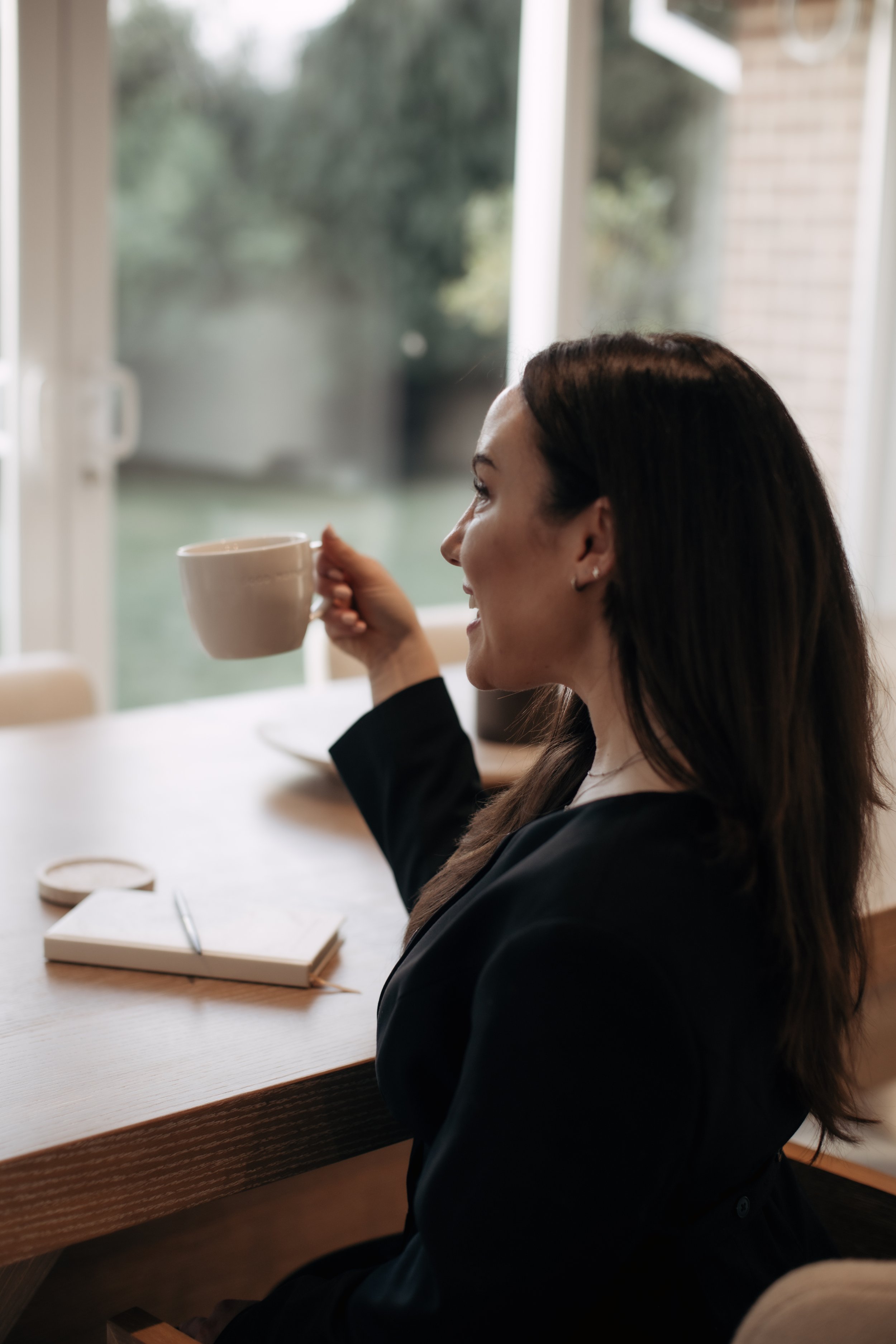 A woman with dark brown hair sitting at a wooden table, holding a white mug and smiling, with a notebook and pen in front of her, near a window with a view of a backyard.