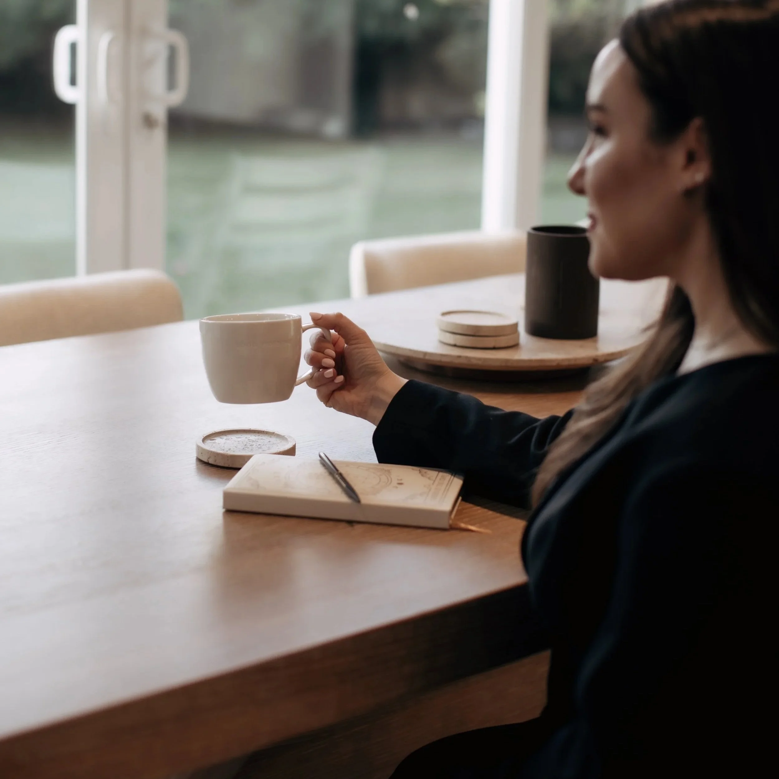 Woman sitting at a table holding a white coffee mug, with a notebook, pen, and coasters on the table, in a bright room with large windows.