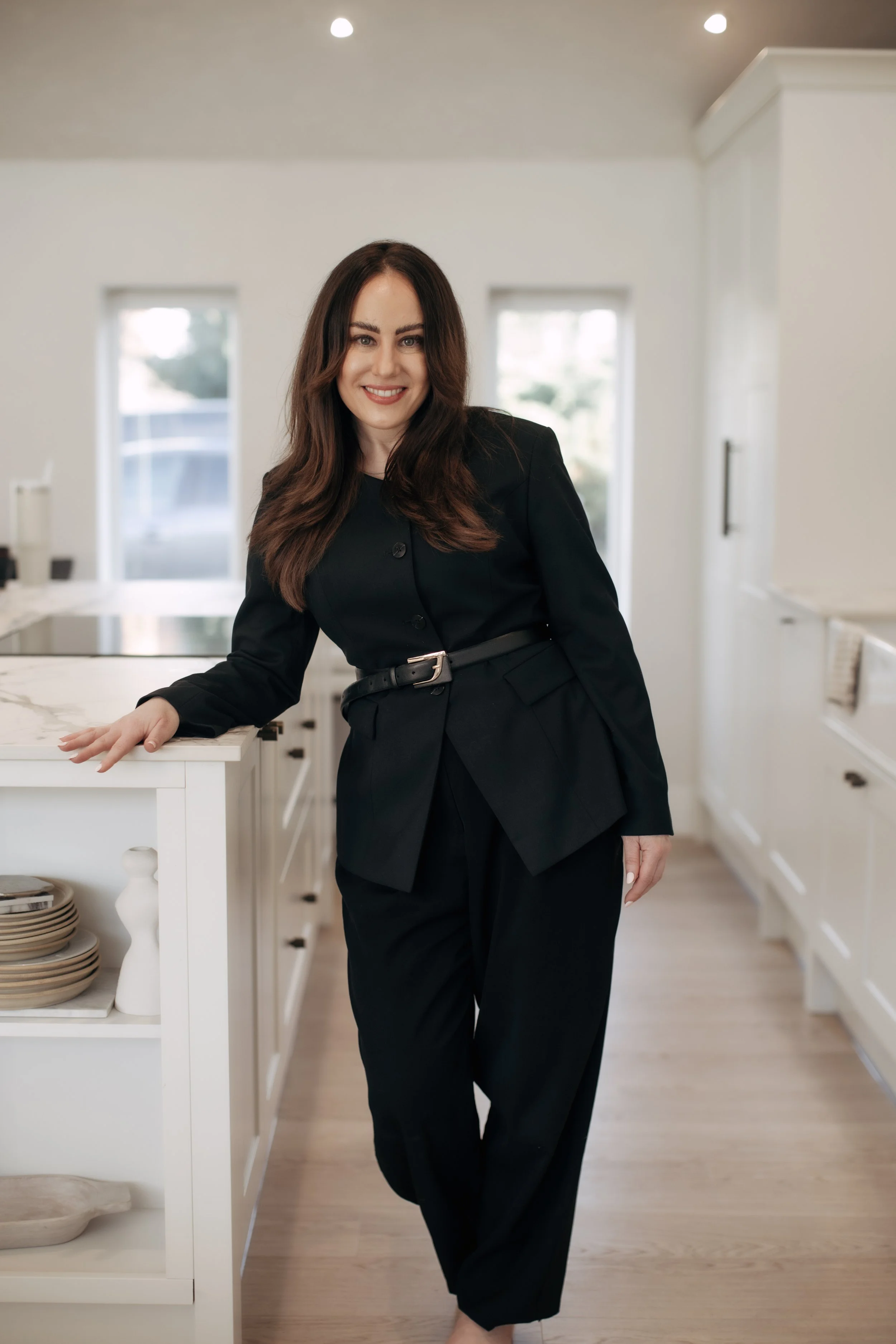 A woman with long brown hair, dressed in a black suit, stands in a modern kitchen leaning on a white marble countertop, smiling at the camera.