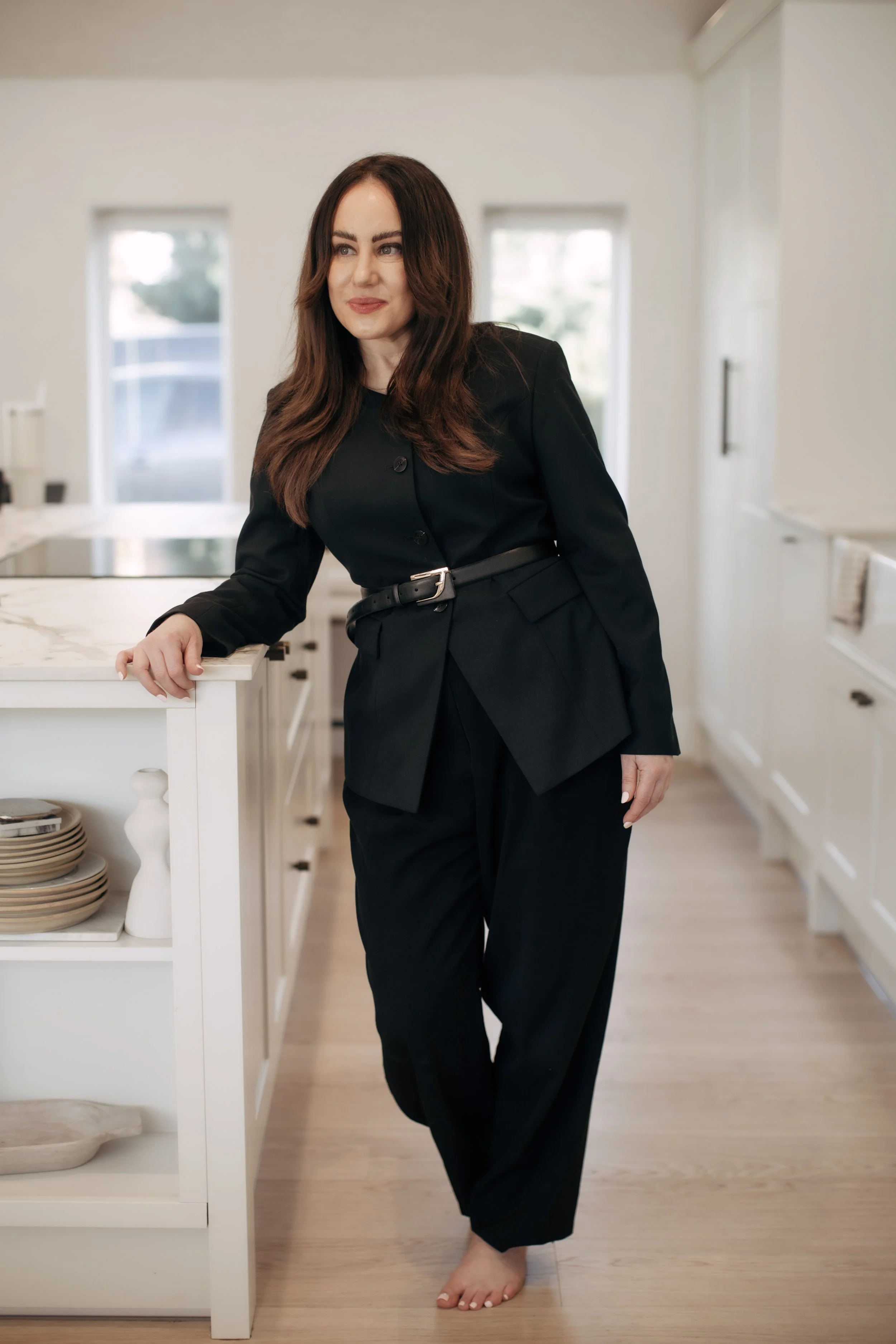 A woman with long brown hair, dressed in a black suit with a belt, standing barefoot in a modern kitchen with white cabinets and wooden floors, leaning on a white kitchen island.