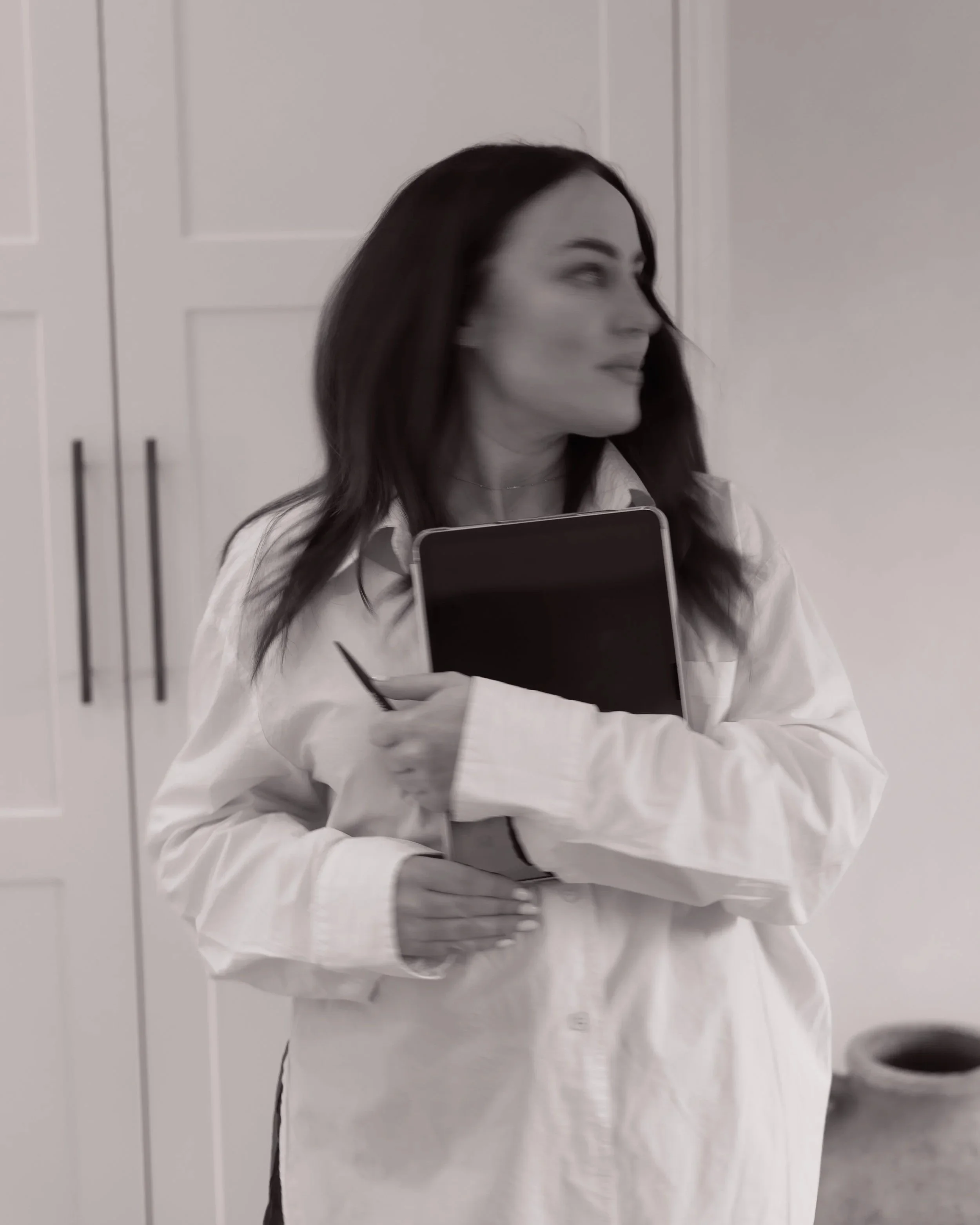 A woman with medium-length dark hair holding a notebook and pen, wearing a white coat, and standing in a room with white cabinets in the background.