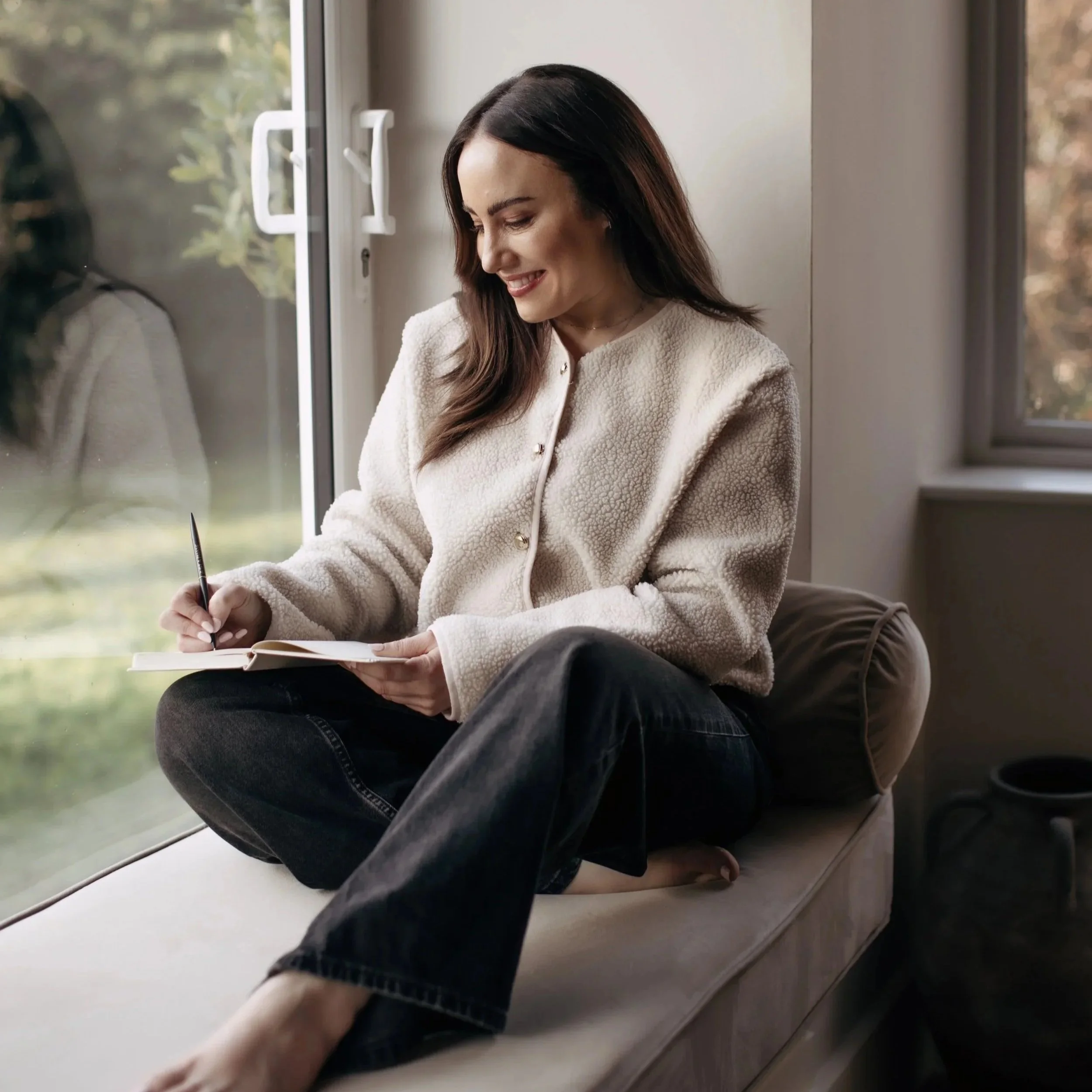 A woman sitting on a window seat, writing in a notebook with a pen, smiling, wearing a cream-colored jacket and black jeans, at home during daytime.