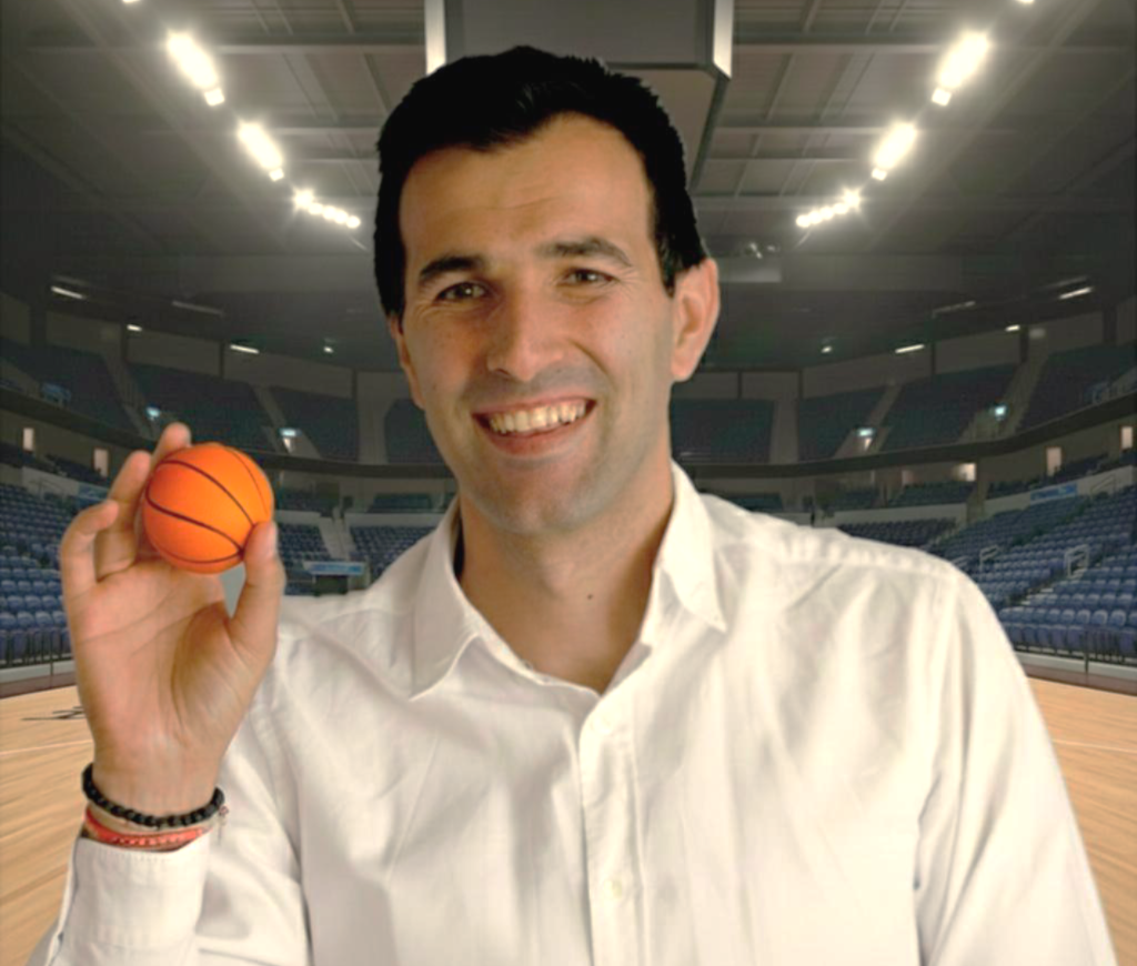 A coach man smiling and holding a small orange basketball in an indoor basketball court.