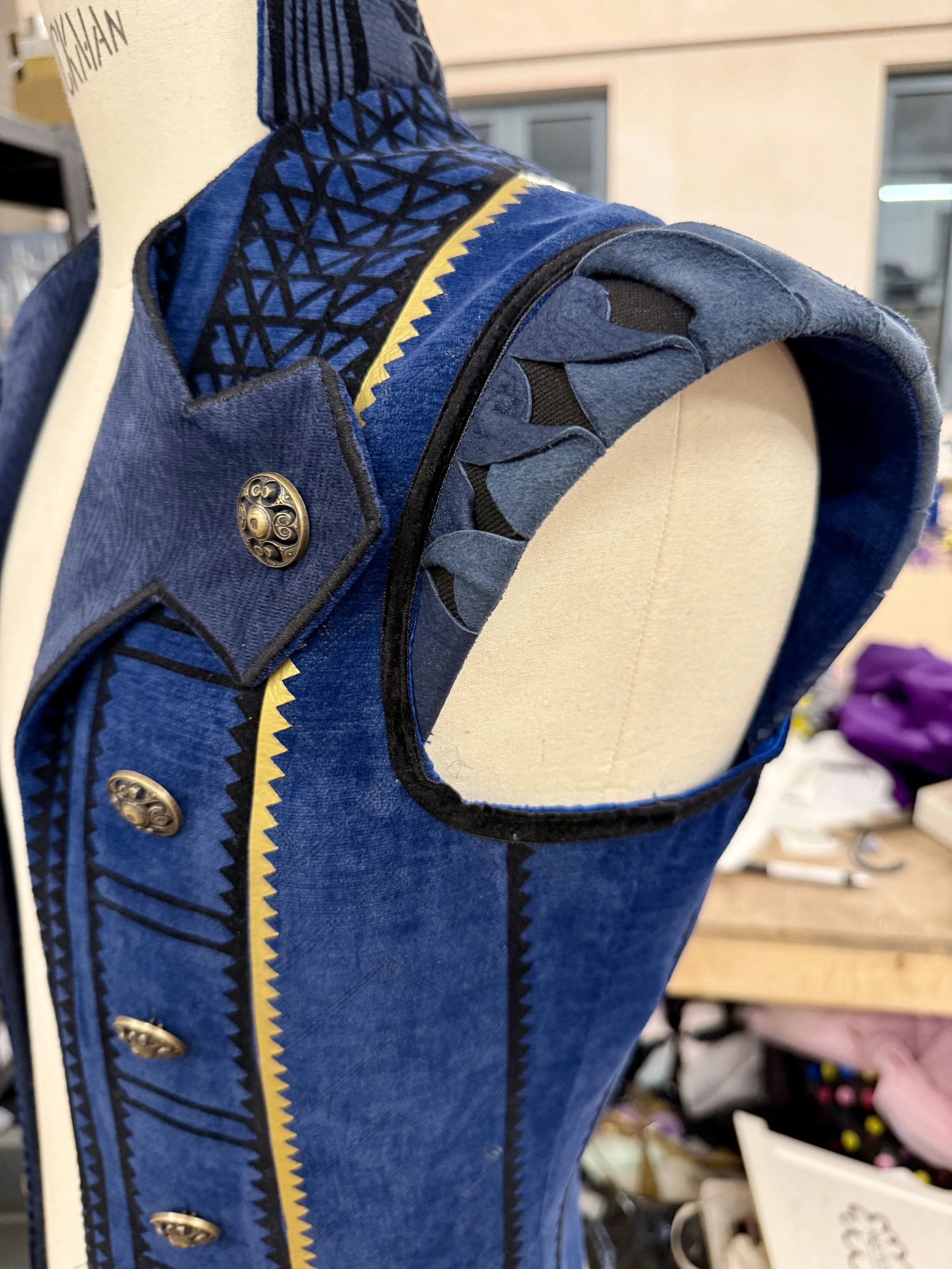 Close-up of a dress form wearing a blue sleeveless garment with detailed black and yellow zigzag patterns, black button embellishments, and fabric ruffled trim around the armhole, in a workshop setting.