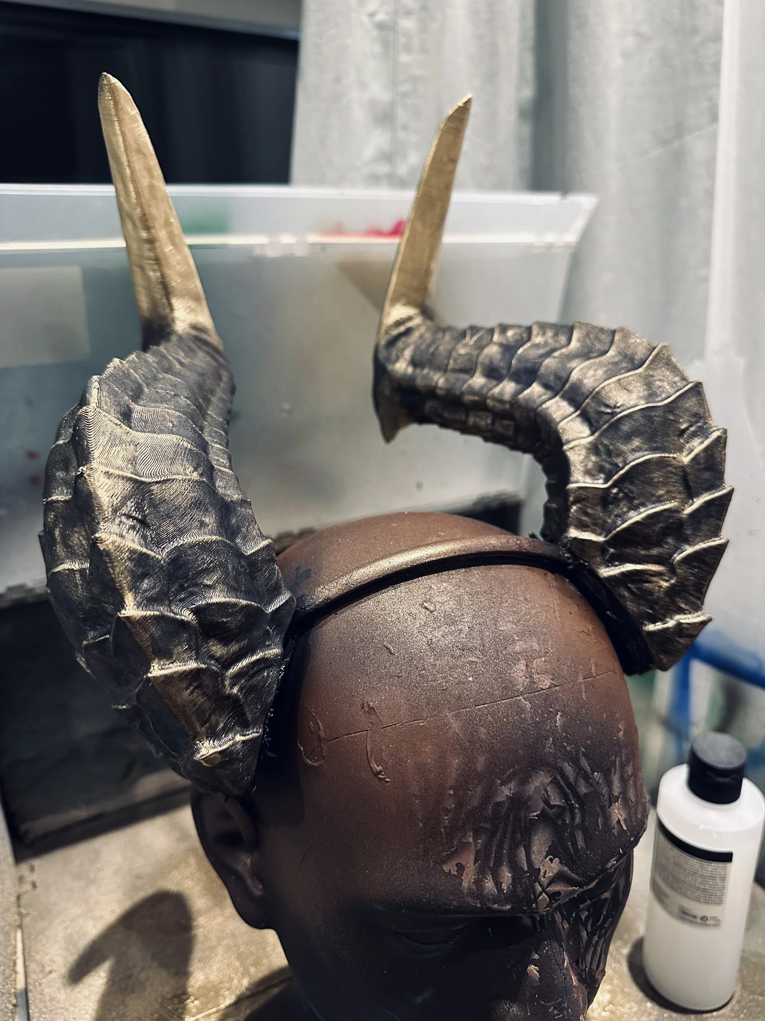 A mannequin head with black skin tone wearing a pair of large, black, textured, curved horns.