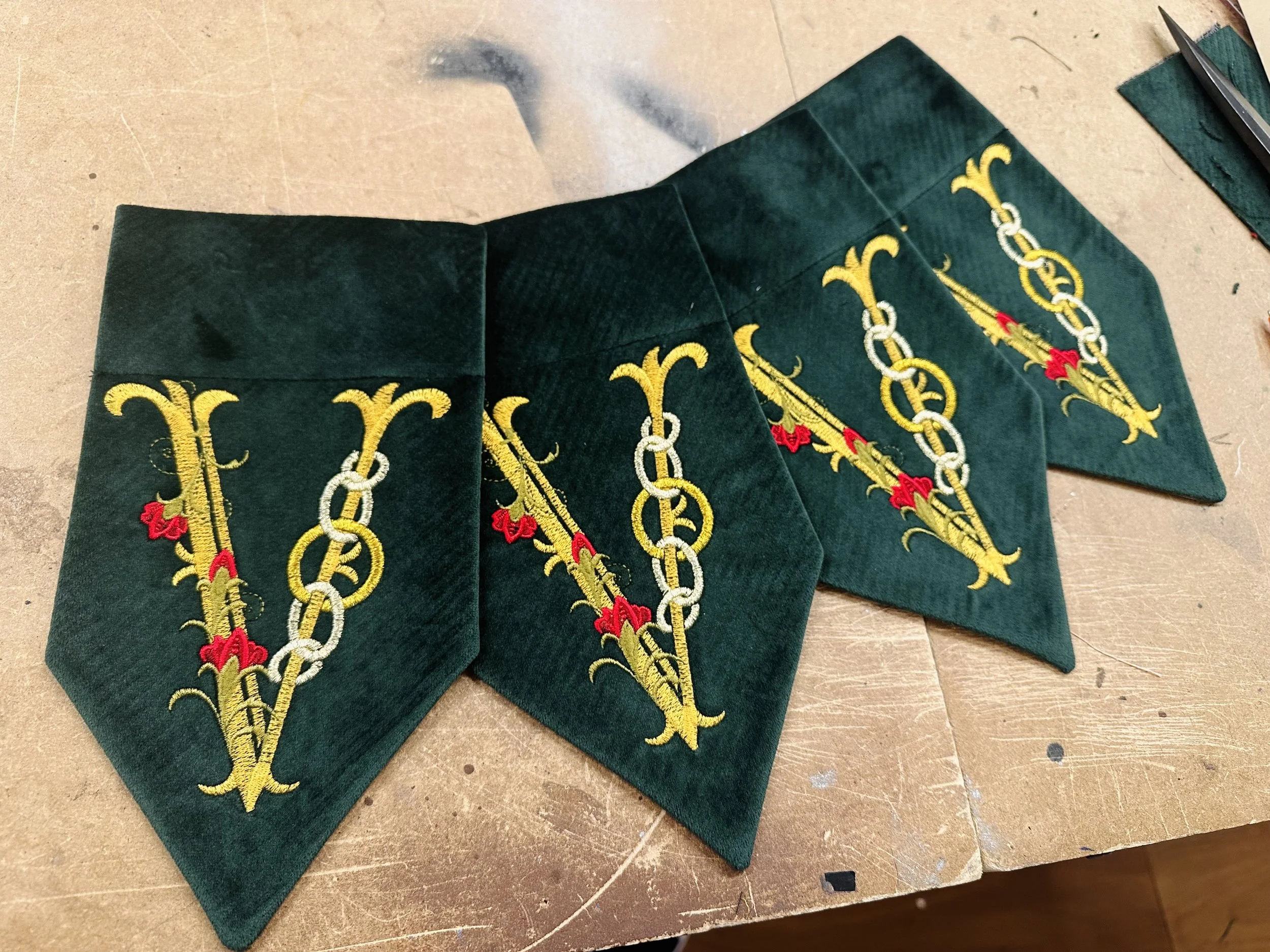 Three green fabric pieces with embroidered yellow, white, and red pattern laid out on a work surface. Each piece has a pointed end, with the pattern consisting of yellow scrolls and chains with red accents.
