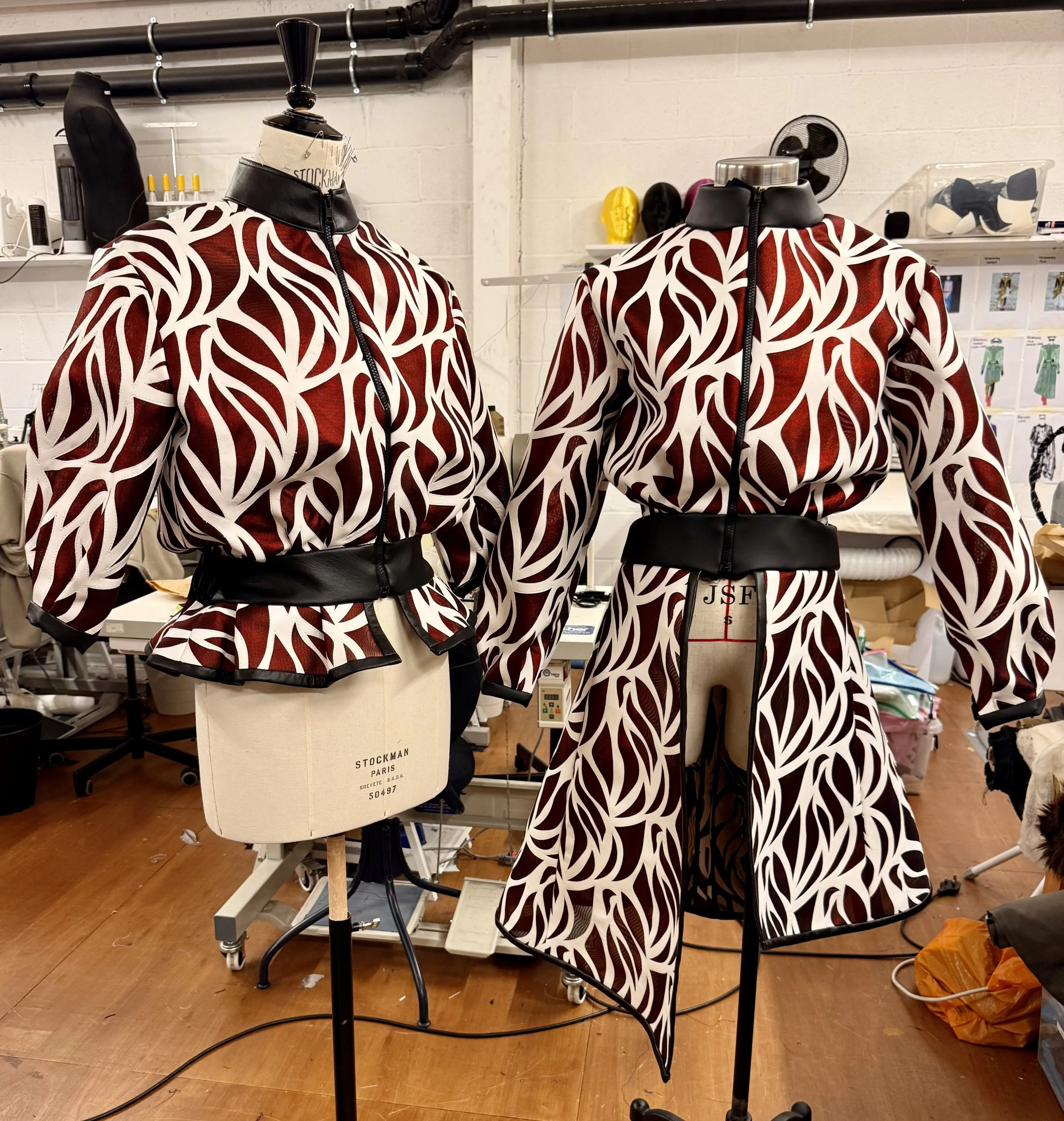 Two mannequins wearing matching red and white patterned jackets in a workspace with sewing equipment and designs in the background.