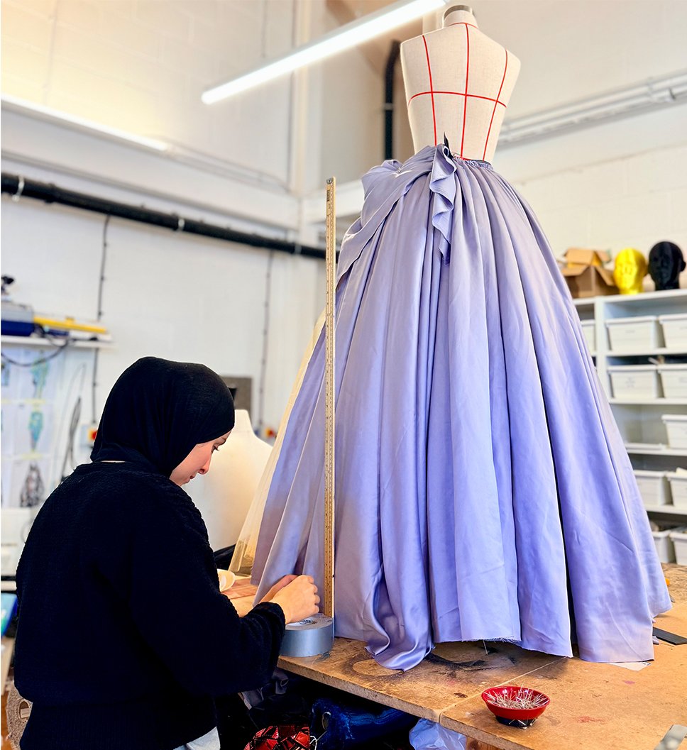 A fashion designer fitting a lavender-colored satin gown on a dress form in a studio.