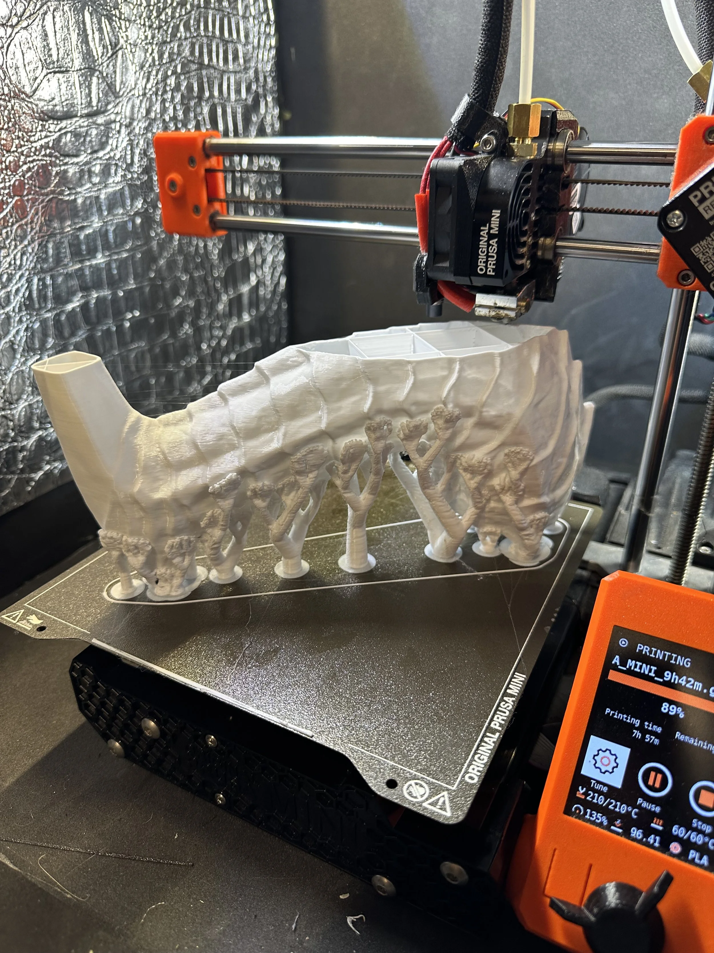 A 3D printer printing a white, hollow, ribbed structure that resembles a section of a bone or coral, positioned on a print bed surrounded by metal and filament components.