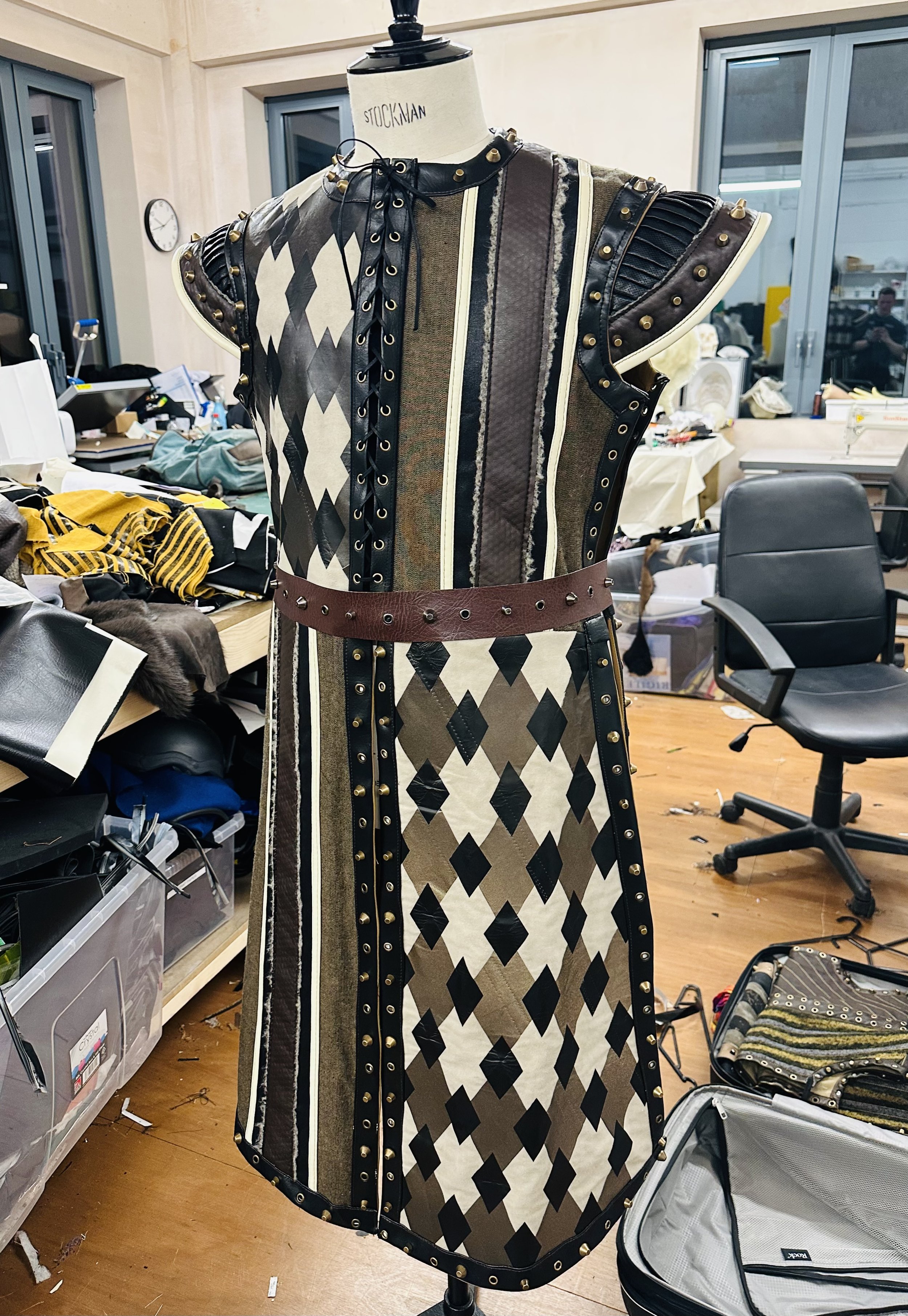 A mannequin wearing a patchwork dress with various patterns and eyelets, set in a cluttered workspace with chairs, boxes, and fabric around.