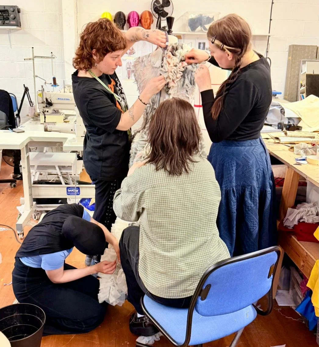 Four people working together on a dress or costume in a workshop. The dress is on a mannequin, and they are sewing or attaching fabric. The workshop has sewing equipment, fabric, and colorful mannequin heads on the shelves in the background.