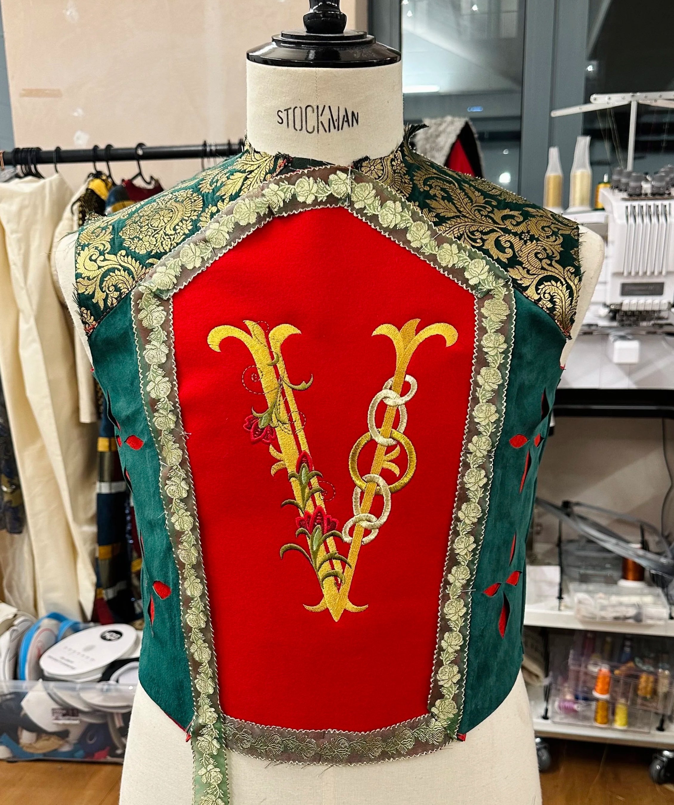 A vintage embroidered vest with gold and red designs on a red background and green sides, displayed on a mannequin with a Stockman label, in a sewing or fashion studio.