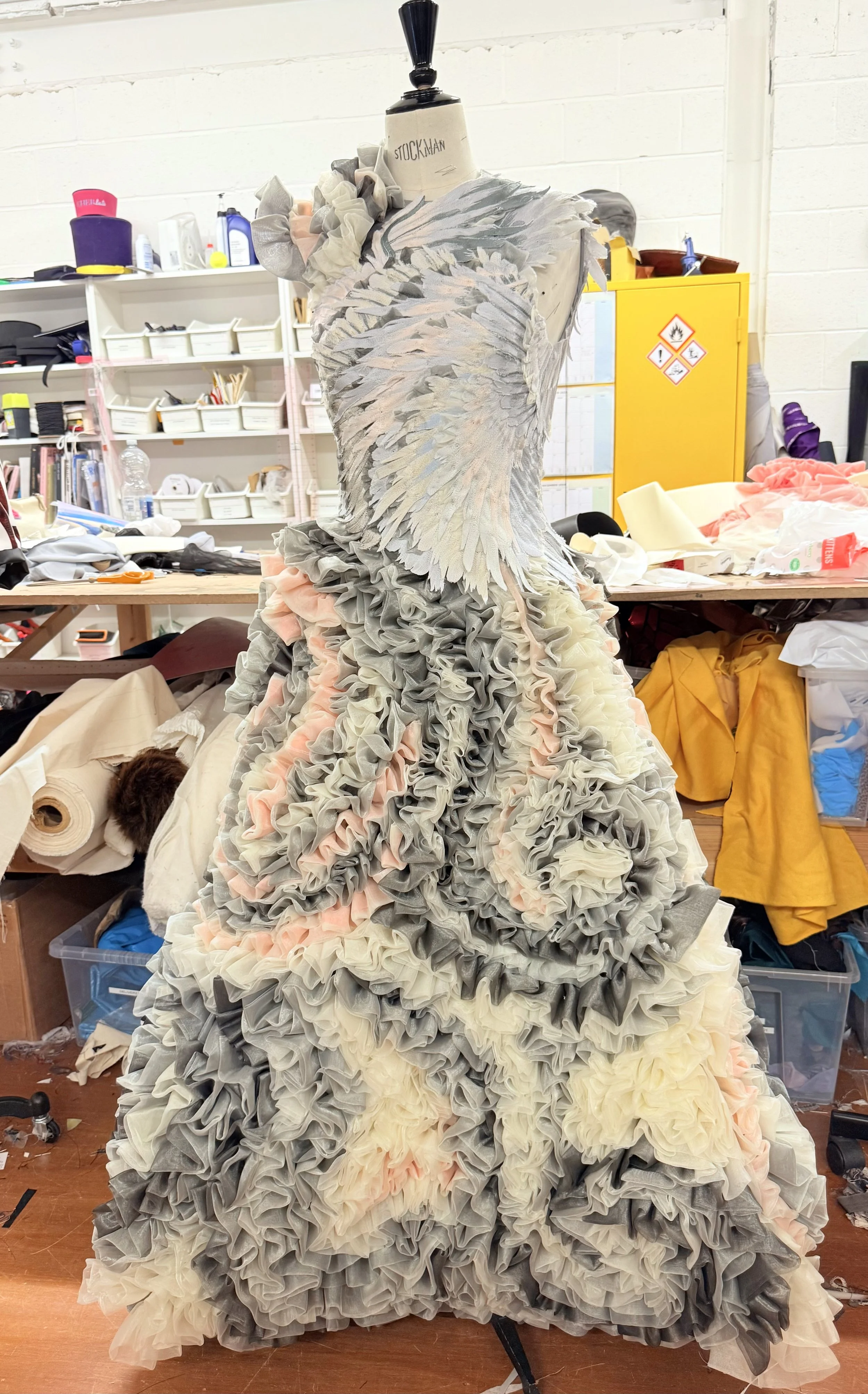 A dress made of layered, ruffled fabric in shades of gray, pink, and cream on a dress form in a workshop or studio