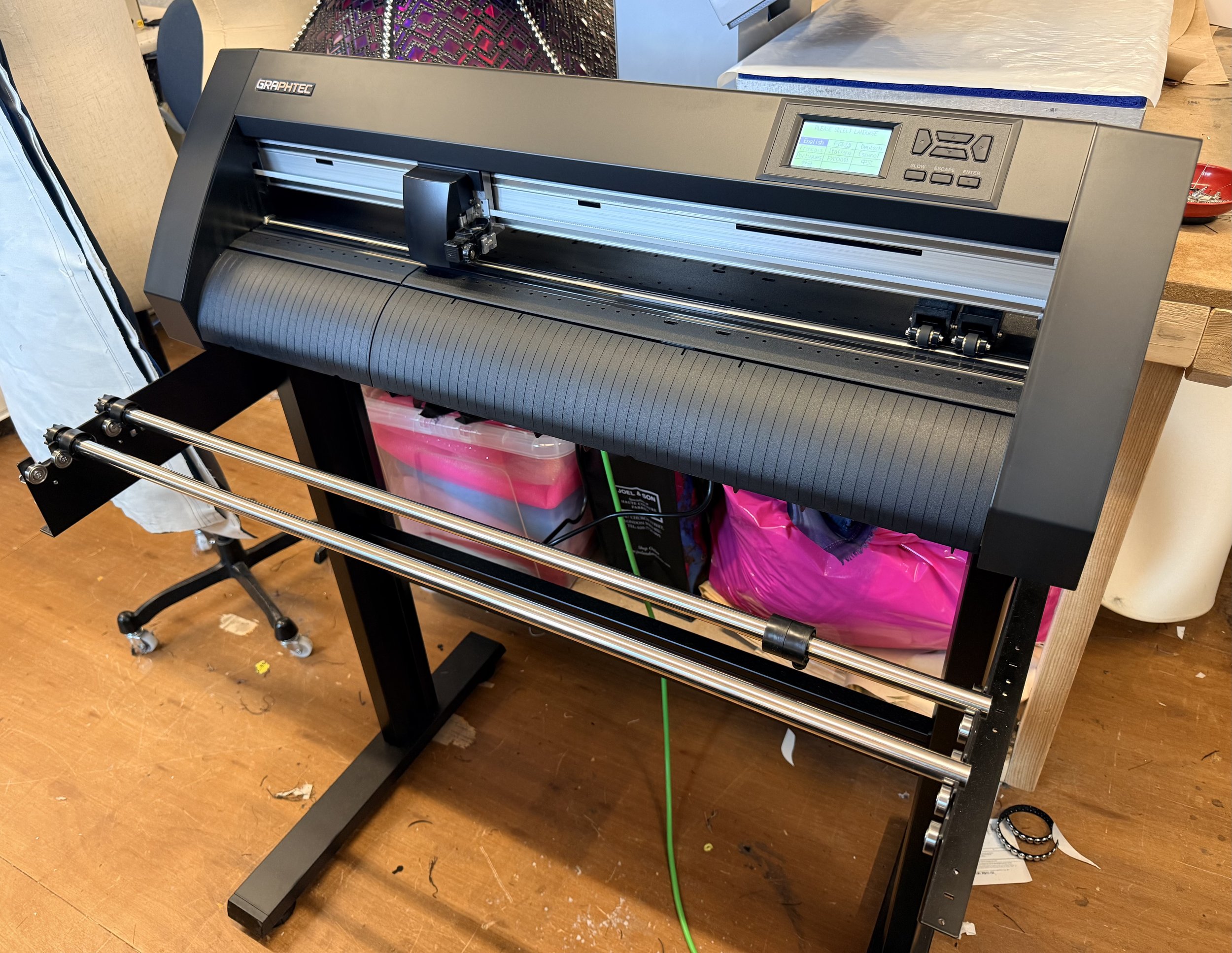 A large wide-format vinyl cutting machine on a black stand with metal support rods, situated on a wooden floor with clutter underneath. The machine is gray and black with a digital control panel on top right. Part of a chair and various items are see