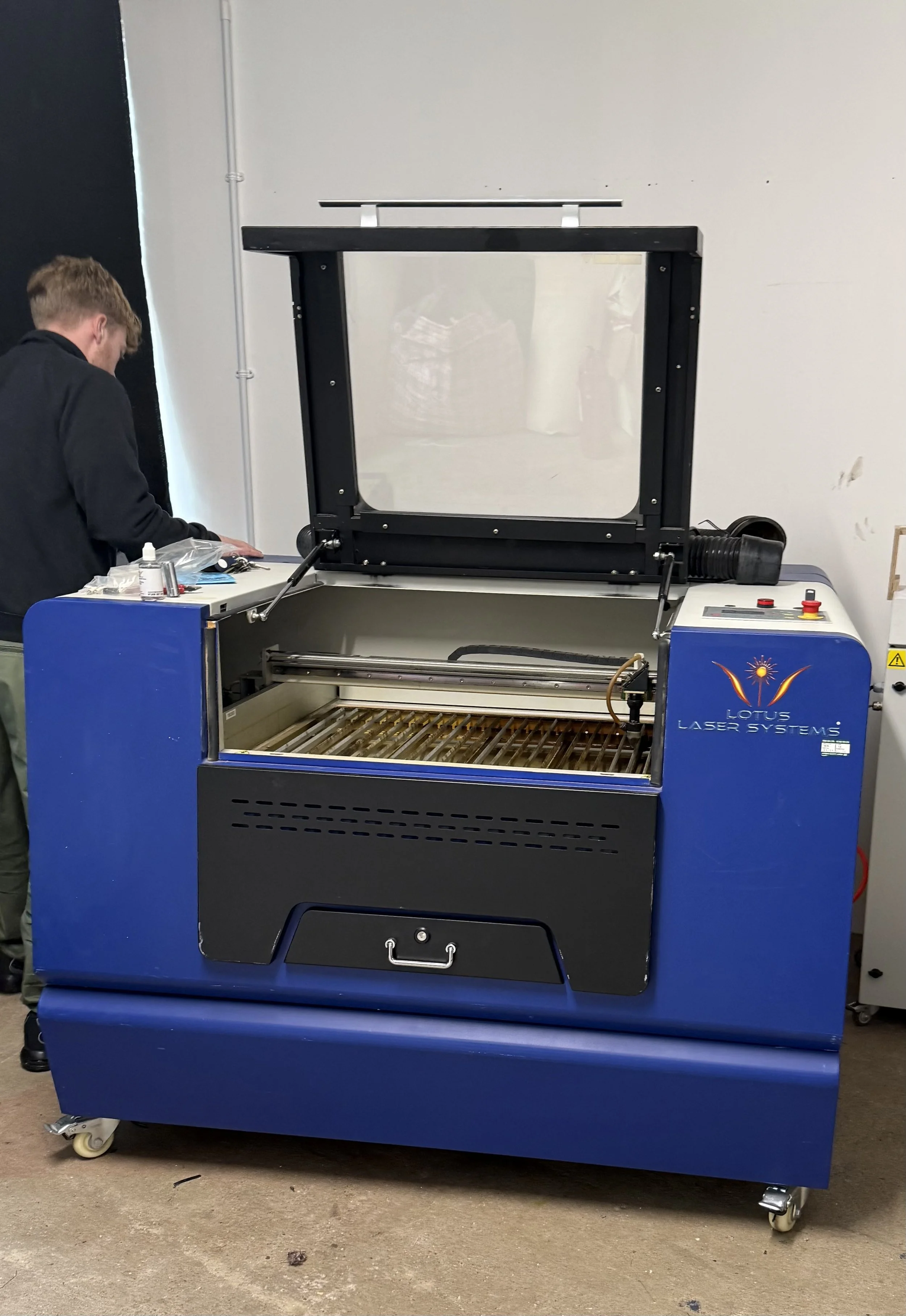 A blue laser cutting machine with an open lid in a workshop or laboratory, with a person working nearby.