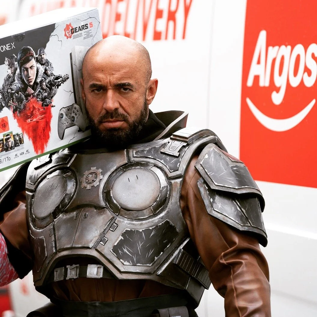 A man with a serious expression wearing a futuristic armored costume, holding a video game box for Gears 5 over his shoulder, inside a store with Argos branding.