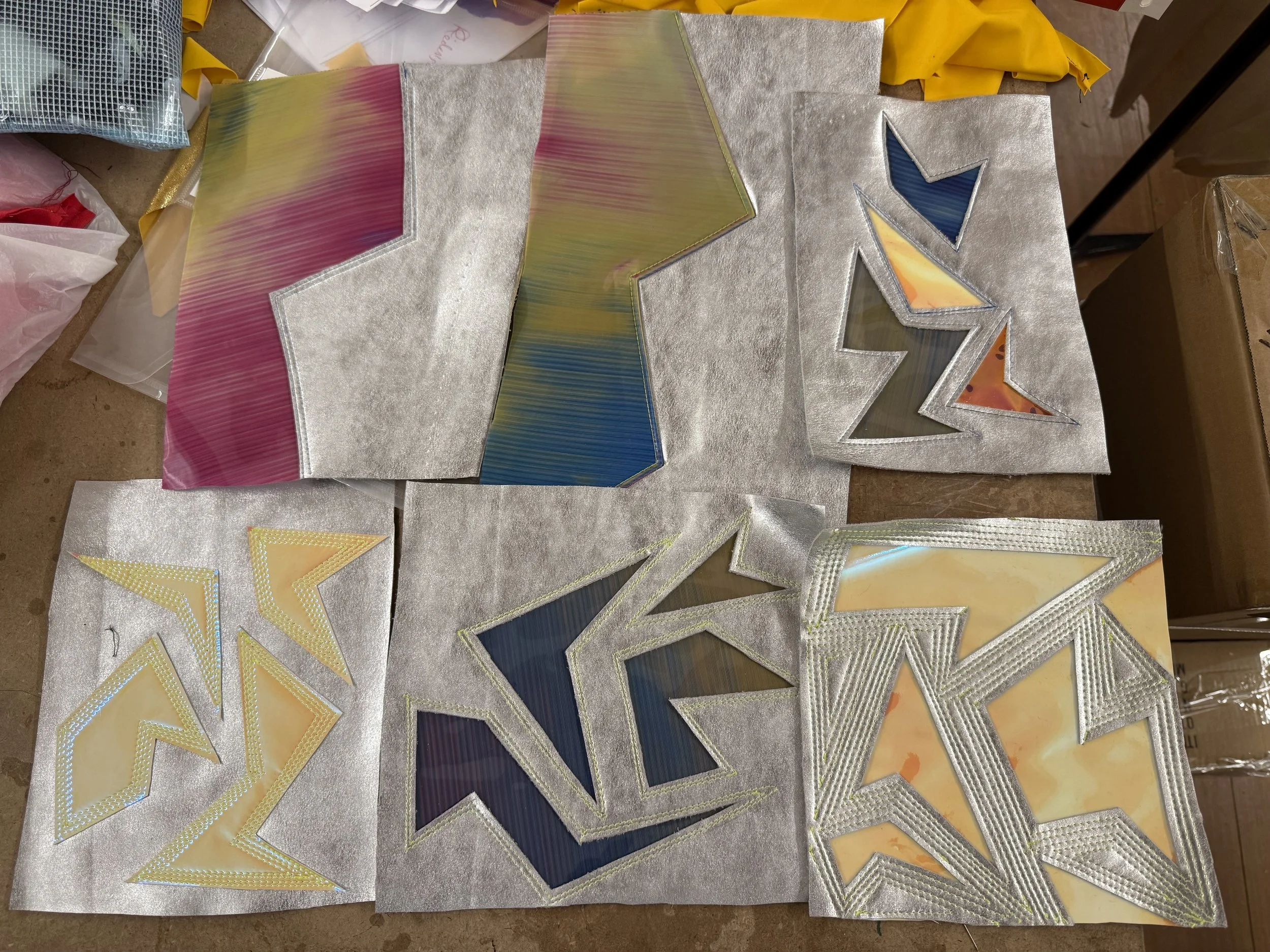 Several stained glass window pieces with colorful, abstract designs and metallic borders, placed on a workspace with paper and fabric around.