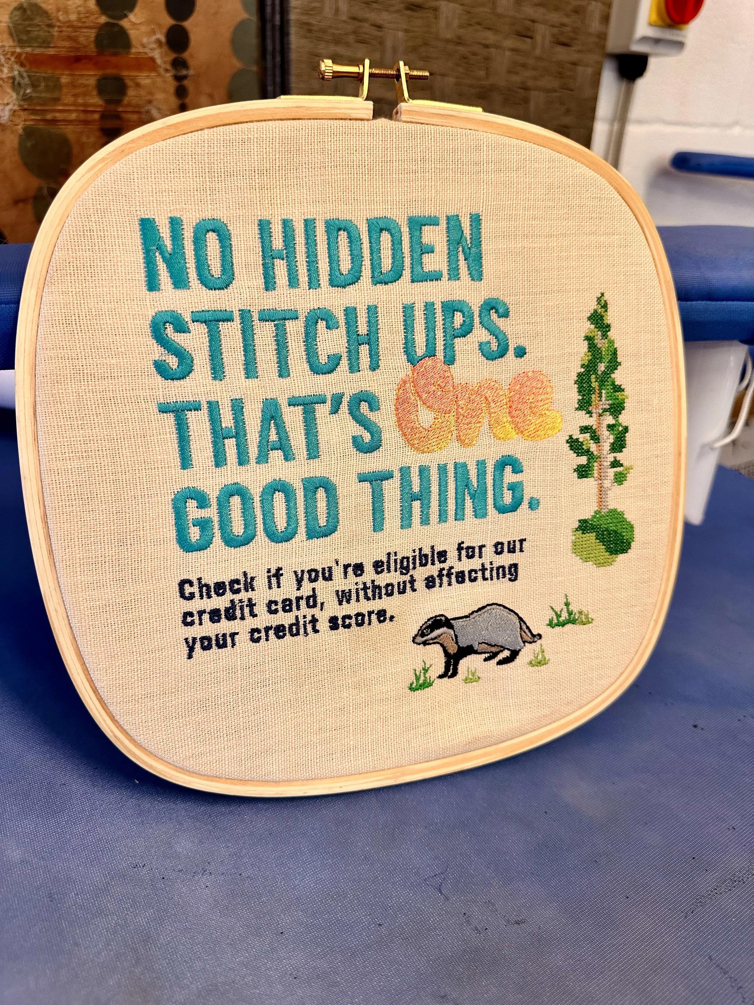 Embroidery hoop with text that reads, "No hidden stitch ups. That's one good thing." and additional text about credit card eligibility, along with embroidered images of a tree, a pile of leaves, and a badger. capital one, custom embroidery, billboard