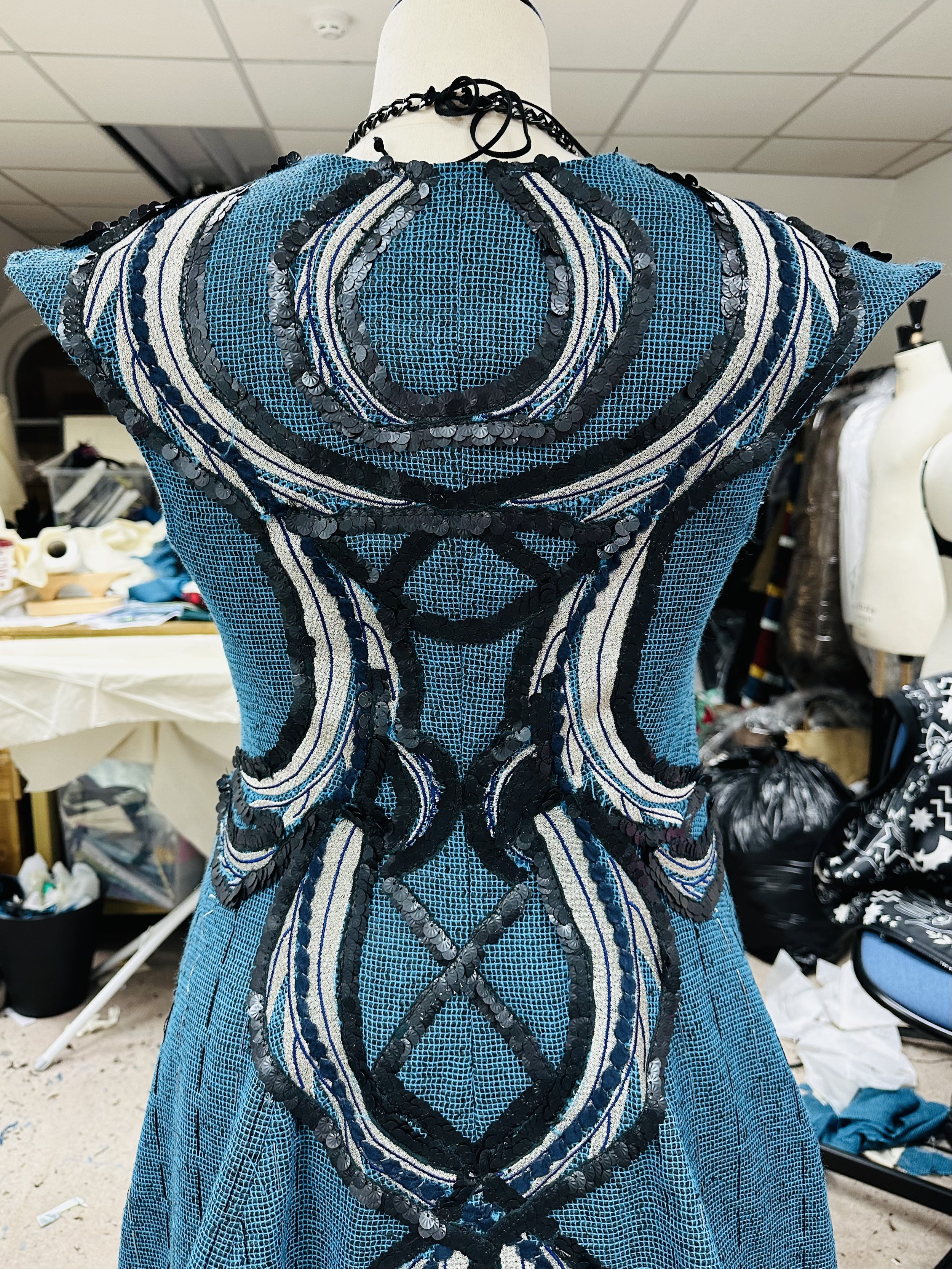 Blue dress with intricate black and white sequin and thread embellishments displayed on a mannequin in a cluttered workshop.