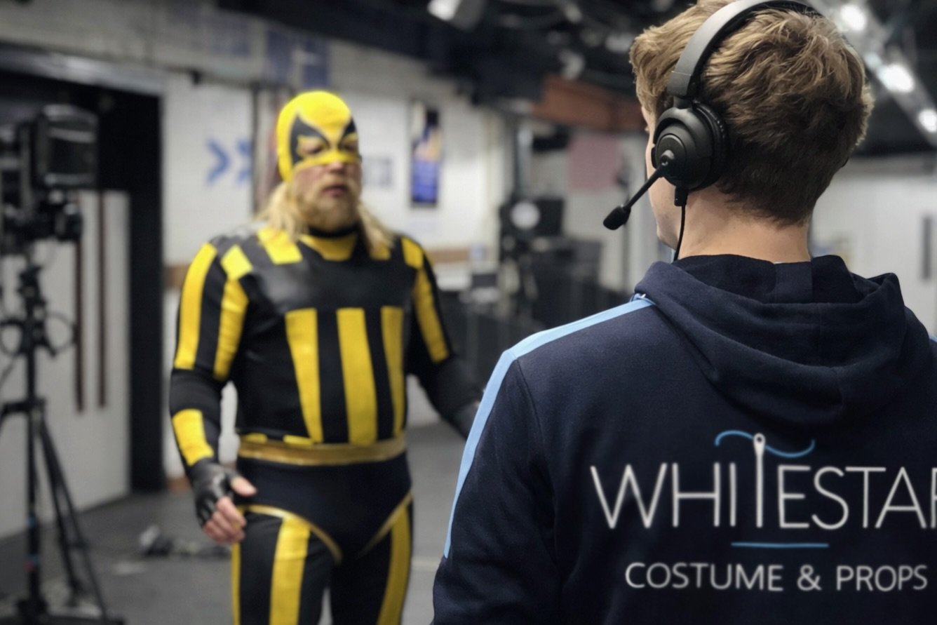 A person wearing a blue hoodie with 'WHITE STAR COSTUME & PROPS' on the back, wearing a headset, is talking to a person in a black and yellow wrestling costume and mask in an indoor setting with cameras and equipment. Michael McIntyre's big show.