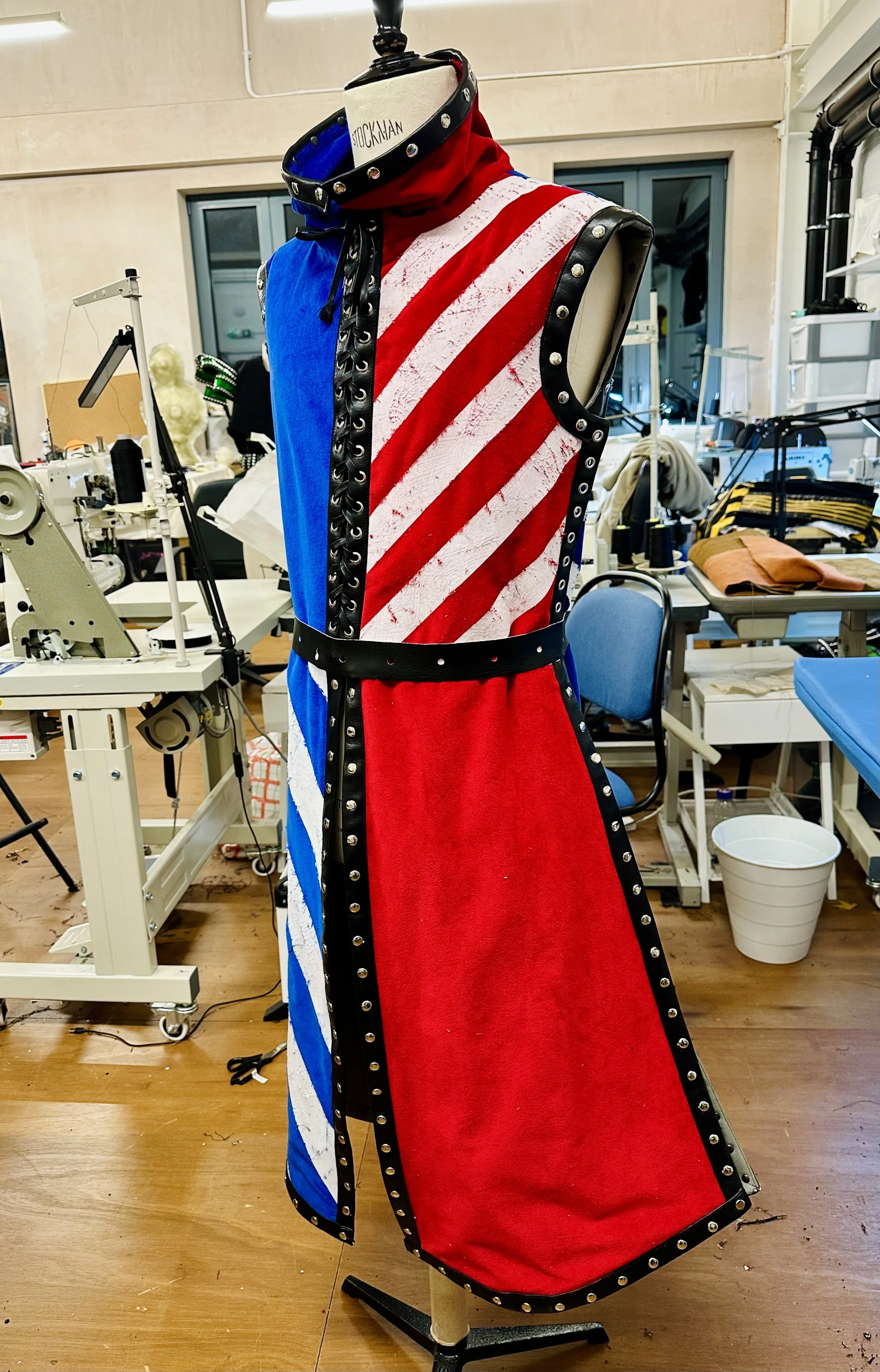 A dress displayed on a mannequin, featuring a design with blue, red, and white striped patterns, with black trim and metal embellishments, in a fashion design or costume workshop.