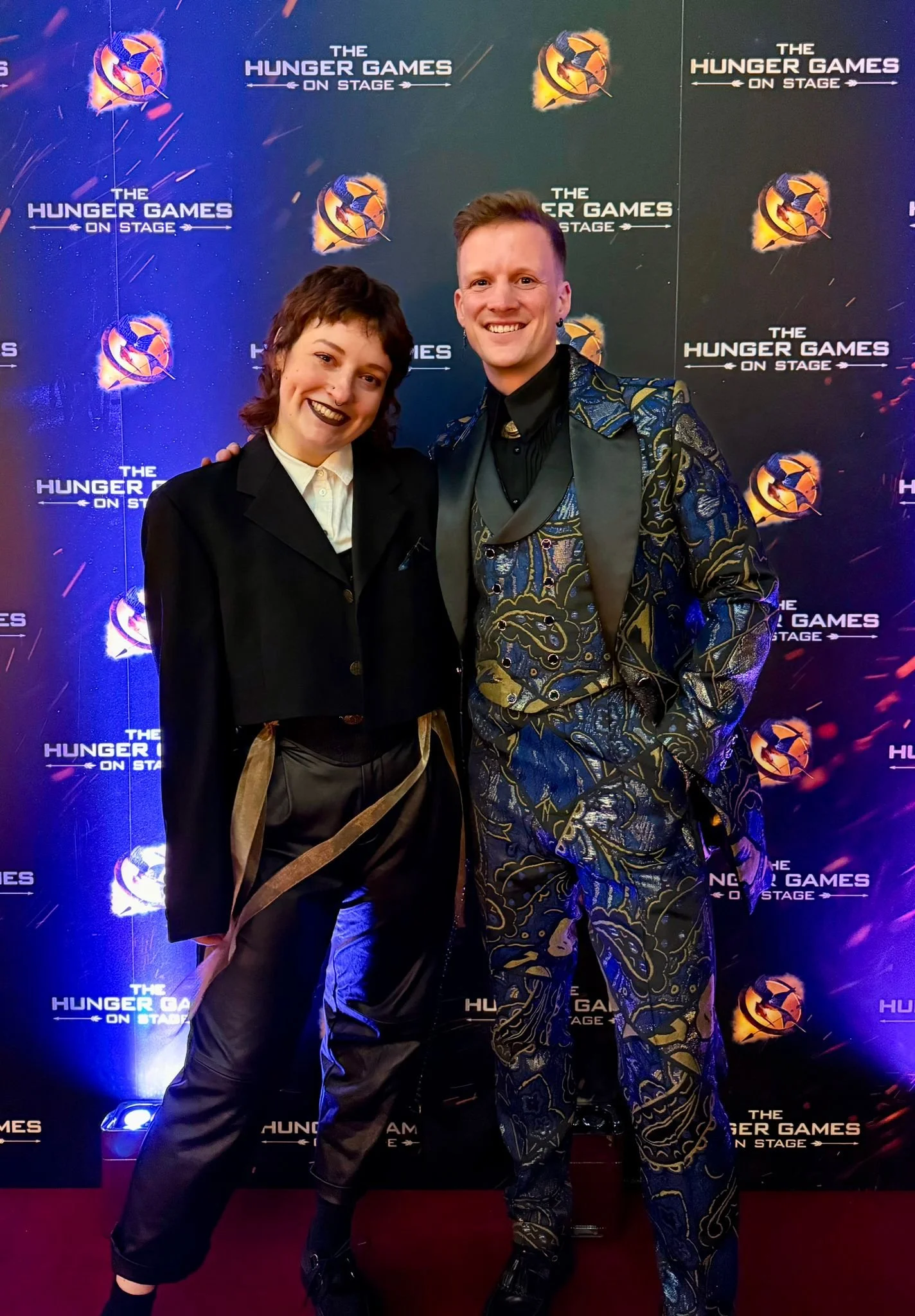 Two people smiling at the camera in front of a backdrop with 'The Hunger Games On Stage' logo, dressed in stylish, formal clothing. Custom outfits made by ourselves at whitestar costume, attending the gala night premiere of the hunger games on stage.