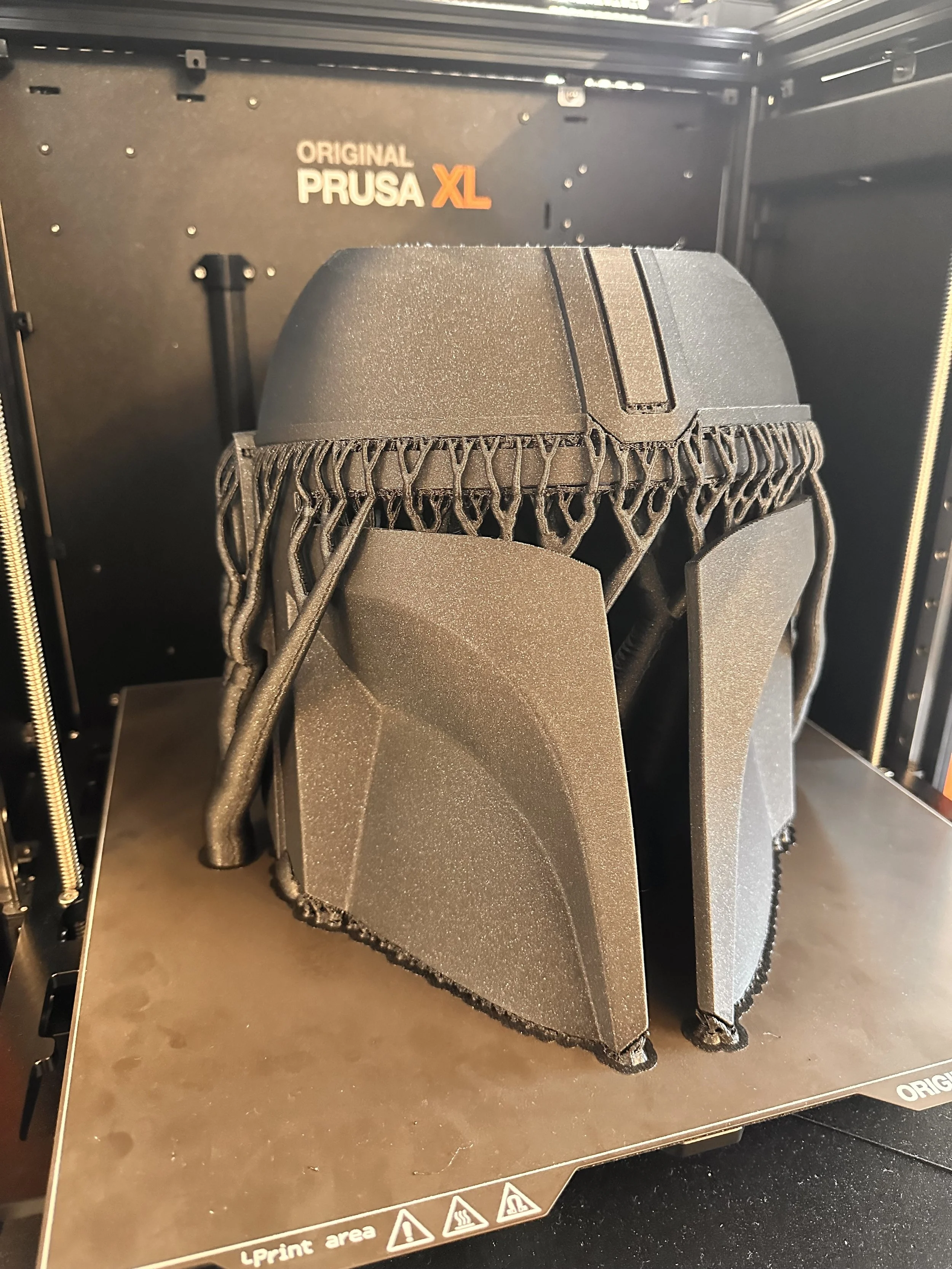 A 3D-printed model of a helmet, mounted inside a 3D printer with the brand 'Original Prusa XL' visible on the printer.