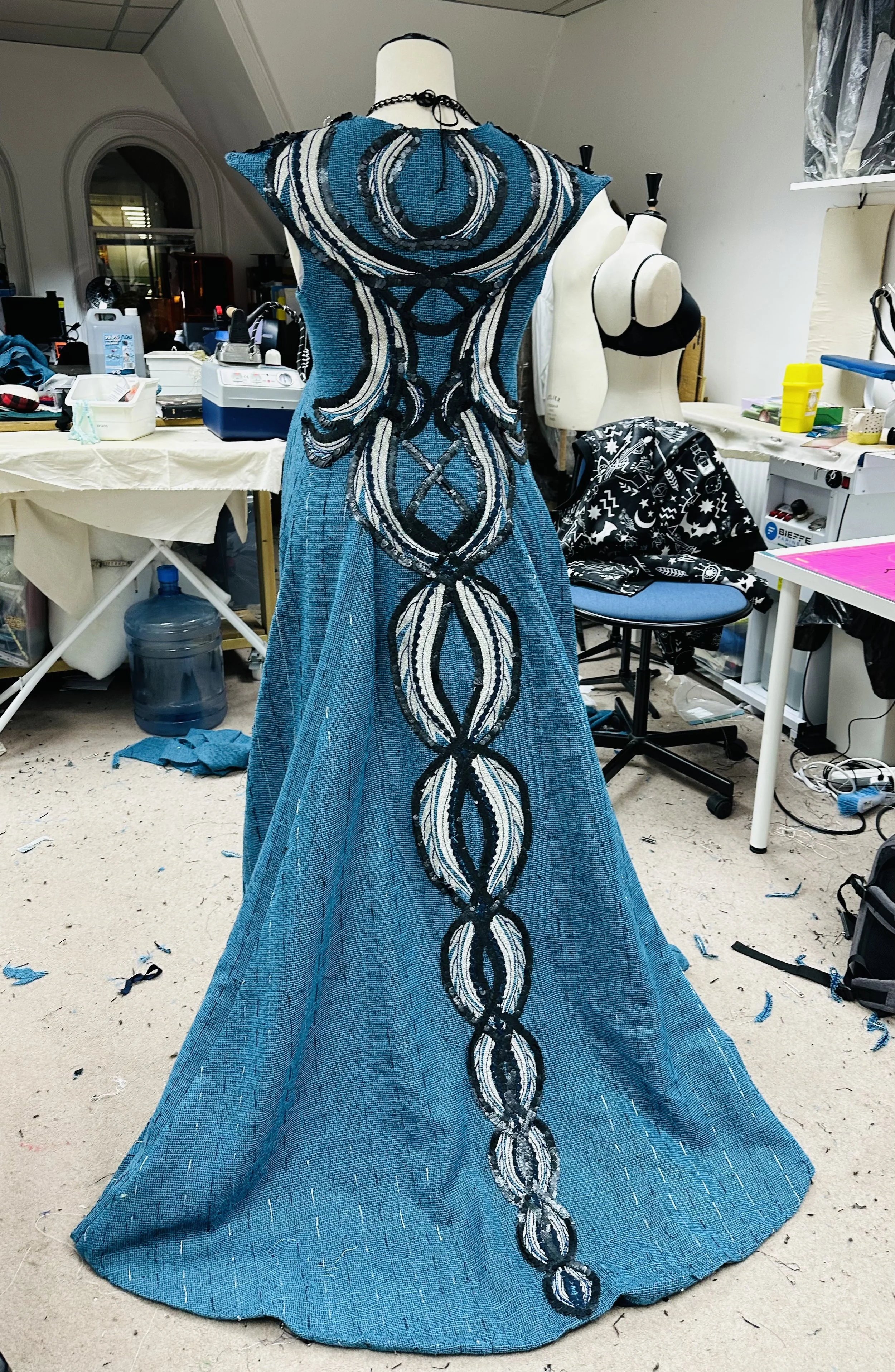 A blue evening gown with black, white, and blue intricate embroidery and beadwork, displayed on a dress form in a sewing workshop.