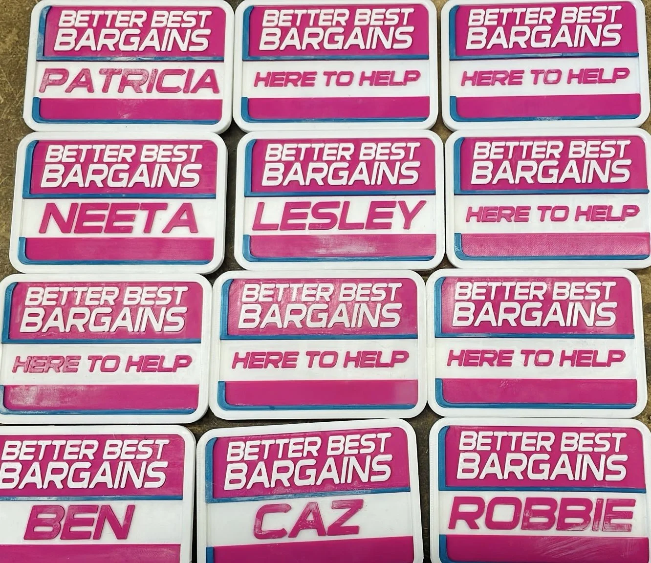 Collection of pink, white, and blue campaign-style signs with pink and white text. The signs display phrases like "BETTER BEST BARGAINS," "HERE TO HELP," and feature names such as "PATRICIA," "NEETA," "LESLLEY," "BEN," "CAZ," and "ROBBIE."