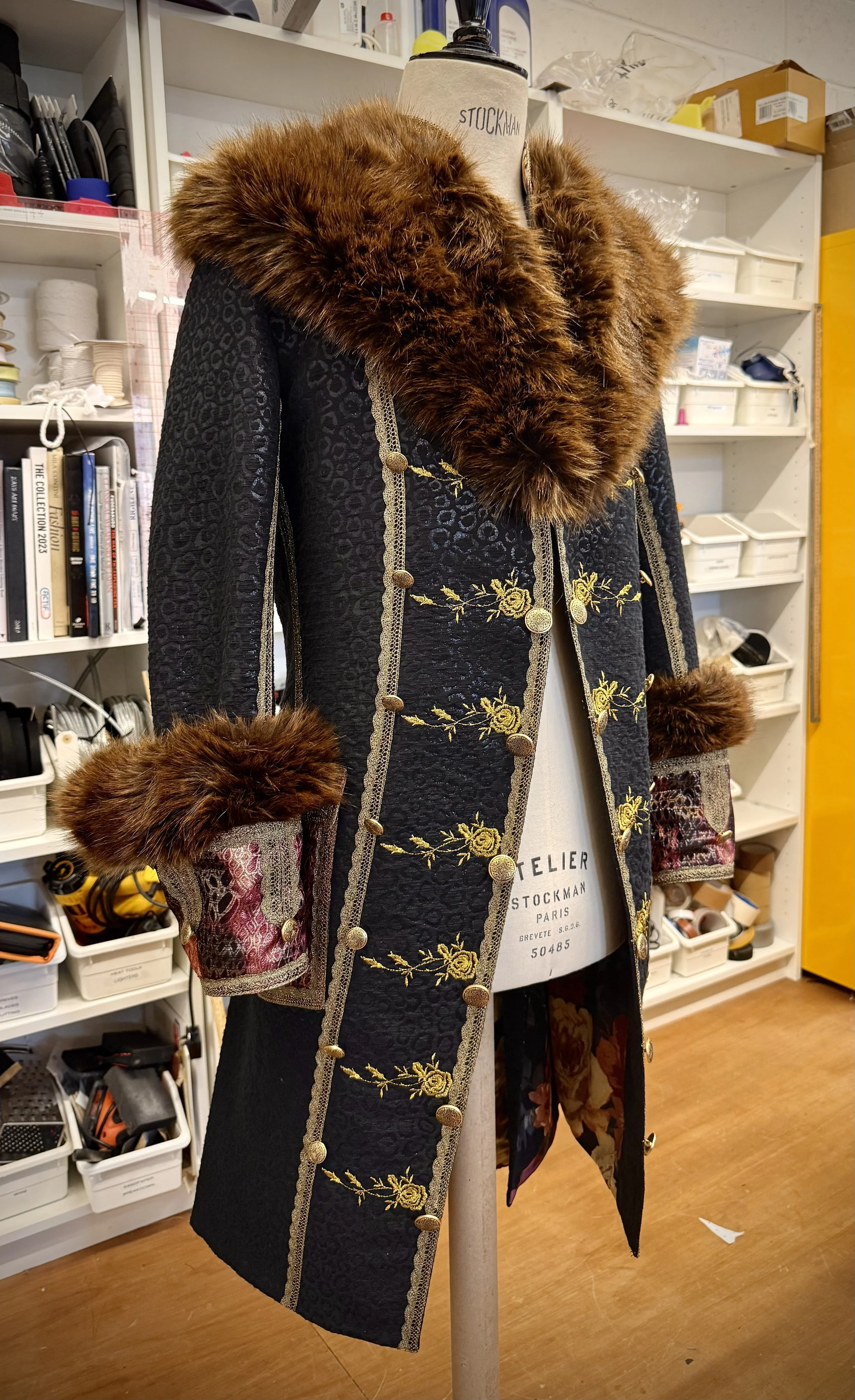 A vintage black coat with floral embroidery and gold buttons, featuring faux fur trim on the collar and cuffs, displayed on a dress form in a cluttered workshop or storage room.