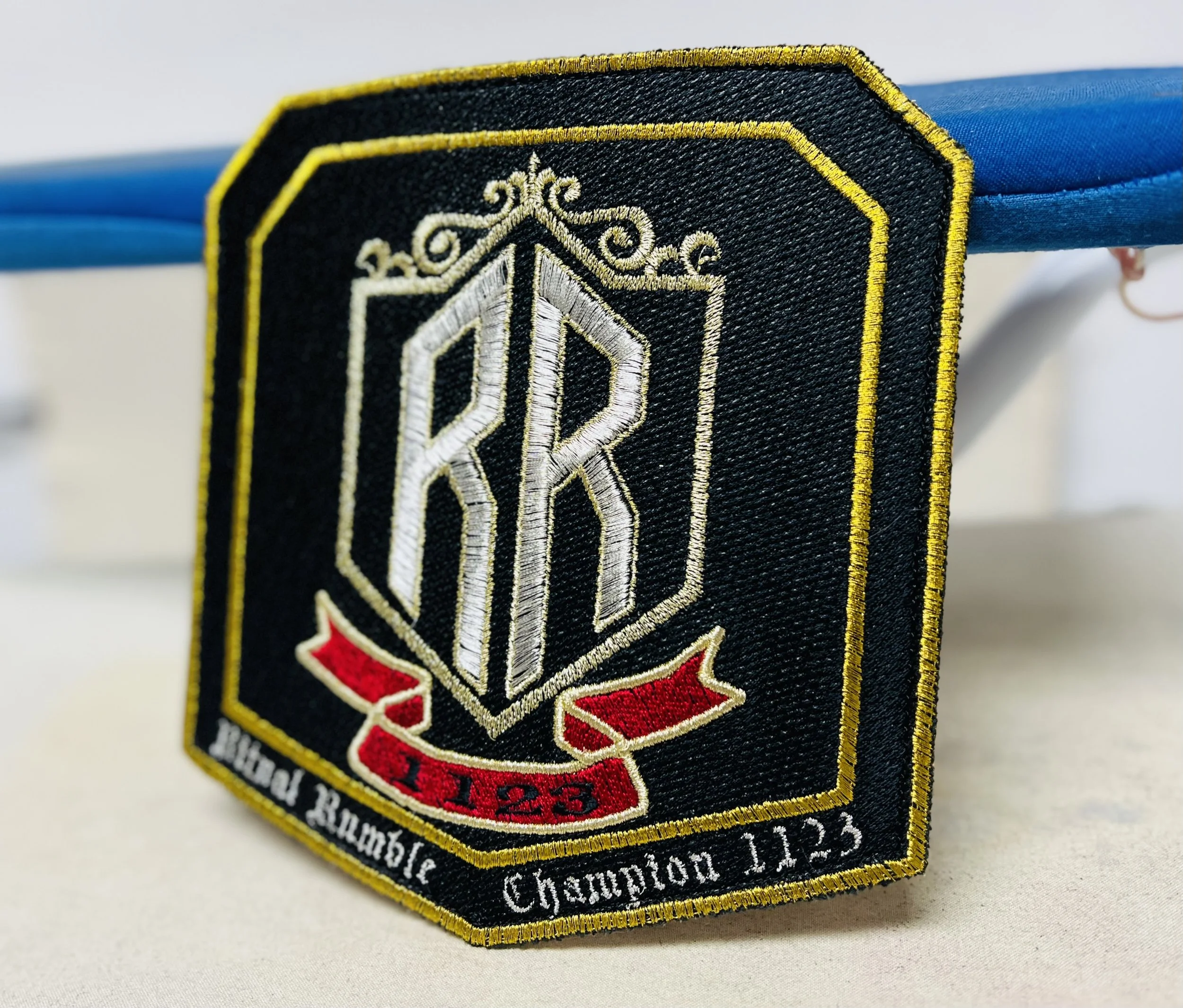 Close-up of a black embroidered patch with gold, white, and red details. The patch features a shield with a stylized monogram and the text "Senior Rambler" and "Championship 11.25." The patch is attached to a blue fabric band.