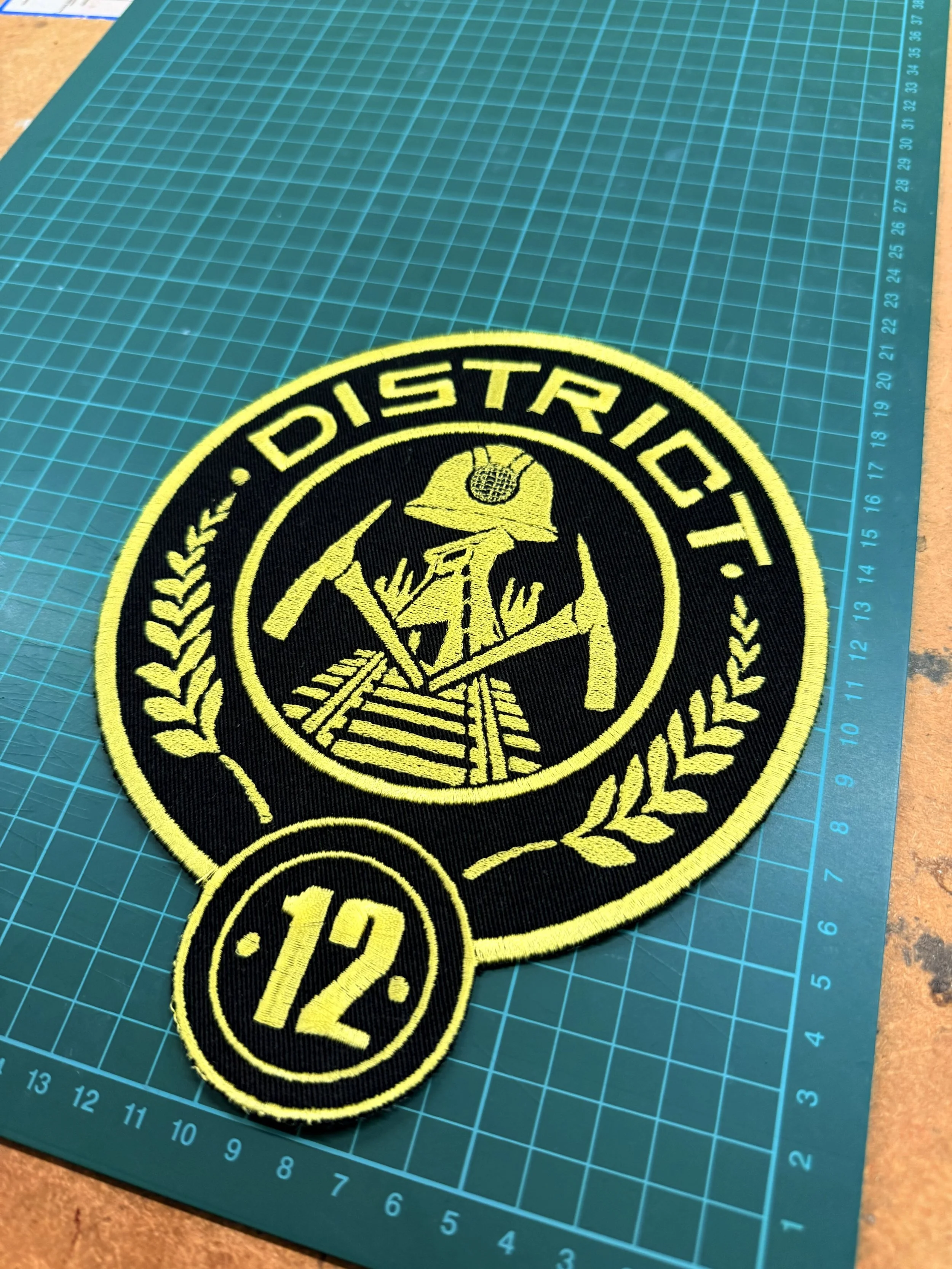 A circular embroidered patch featuring a yellow firefighter skeleton holding an axe, with the text 'DISPATCH' around the top and the number '12' in a smaller circle at the bottom, on a teal cutting mat.