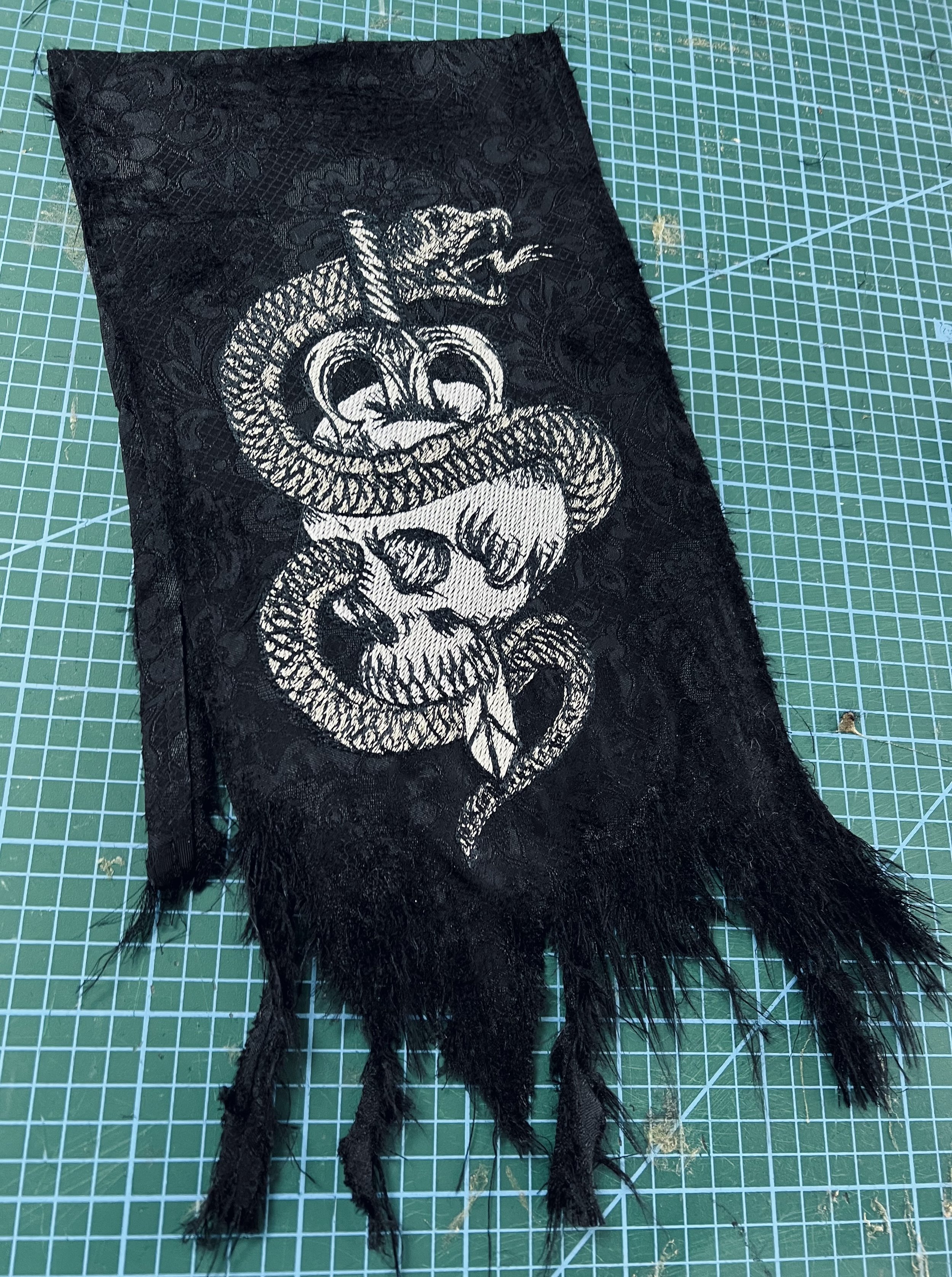 A black fabric patch with a white embroidered snake coiled around a human skull and a snake head above, featuring fringed edges, placed on a green cutting mat with grid lines.