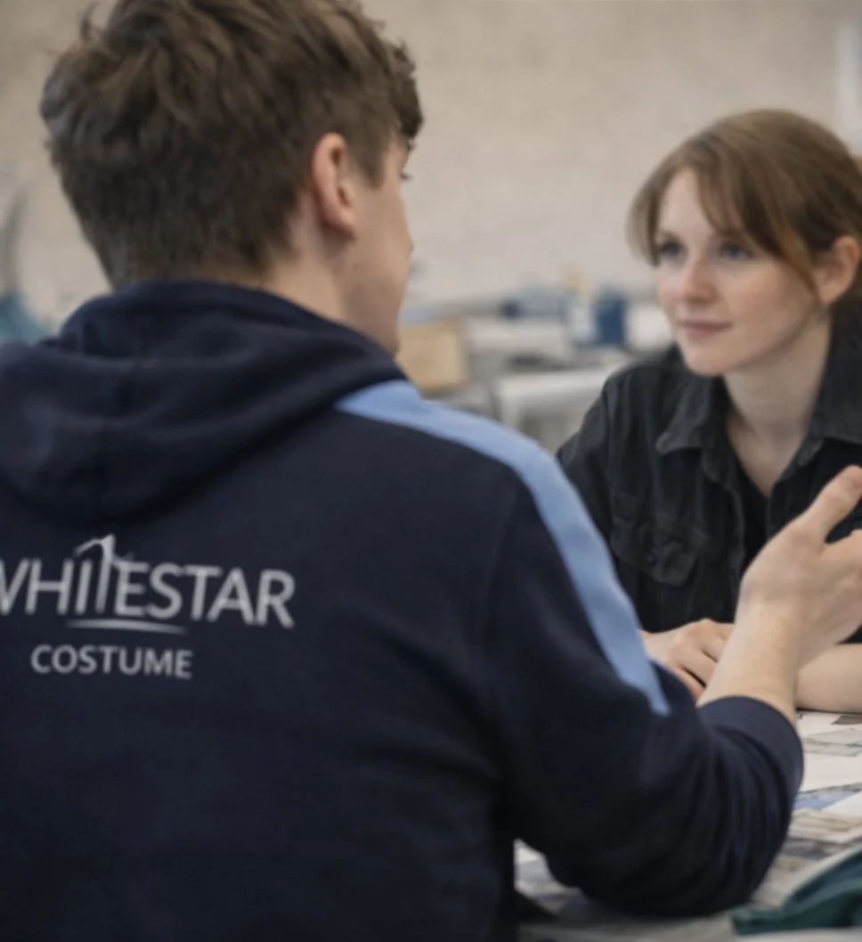 A man and woman are sitting across from each other, engaged in conversation or perhaps an interview. The man has curly hair and is wearing a dark hoodie with the text 'WHITESTAR COSTUME' on the back. The woman has short red hair and is smiling while looking at the man.