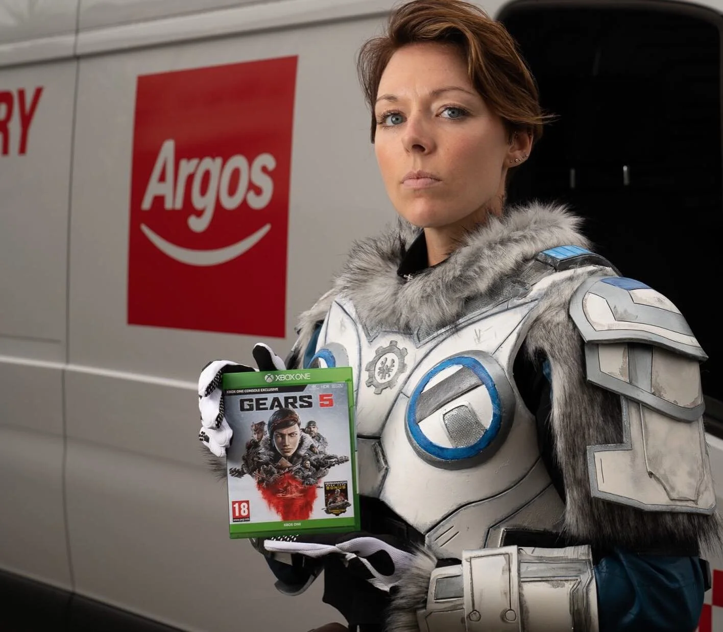 Woman dressed as a futuristic soldier holding a Gears 5 Xbox One game case in front of a vehicle with an Argos sign.