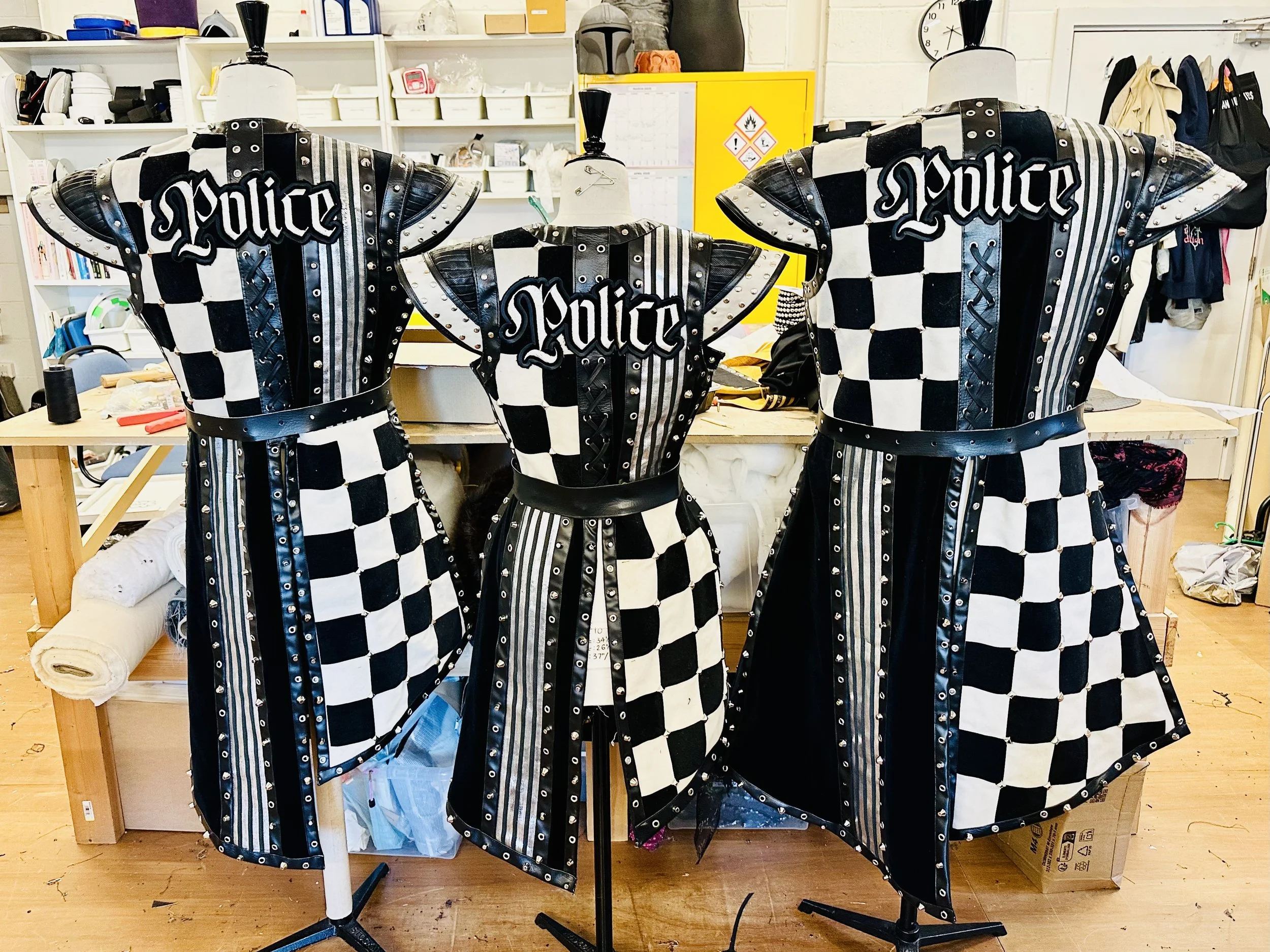 Three mannequins wearing police-themed costumes with black and white checkered patterns, adorned with a large 'Police' patch on the back, placed in a room with shelves, boxes, and miscellaneous items in the background. A knight's tale musical