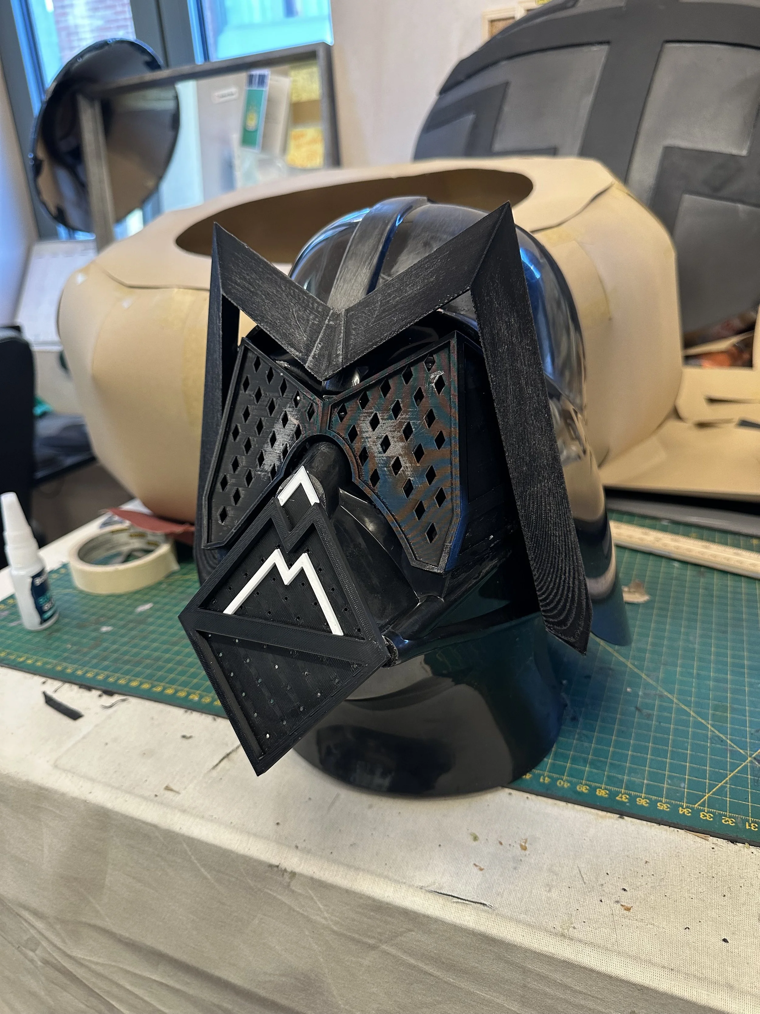 A black and gray sci-fi helmet with angular designs and a geometric faceplate, placed on a green cutting mat on a worktable.