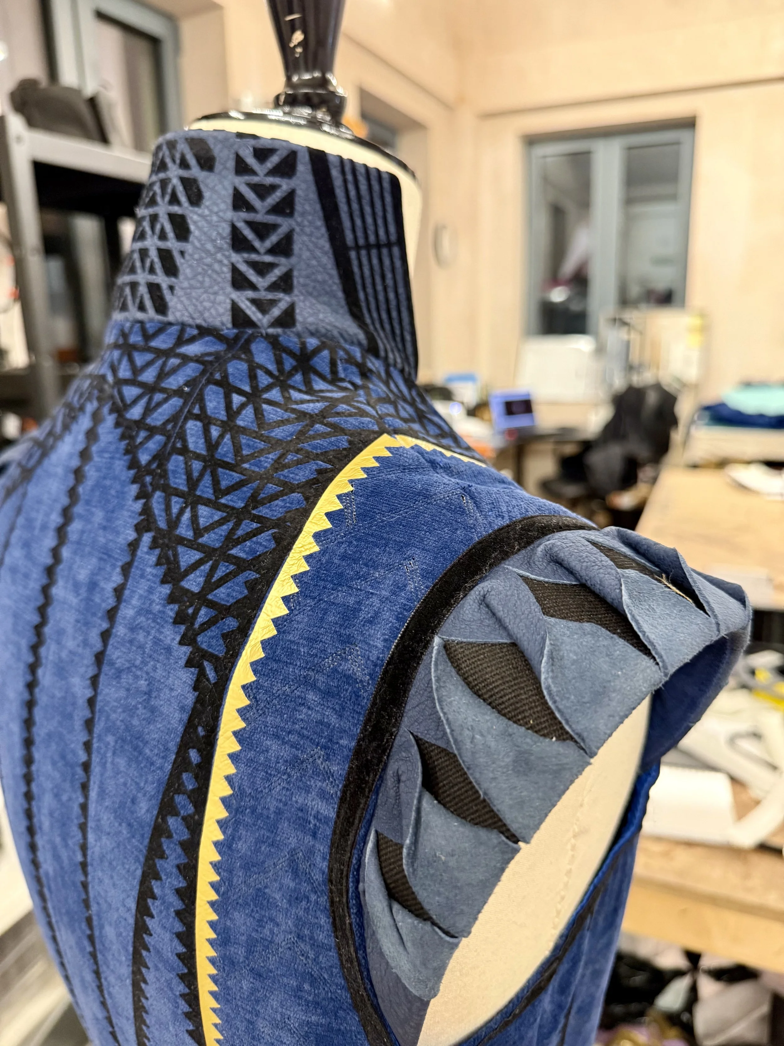 Close-up of a blue and black padded martial arts uniform with textured patterns and a padded shoulder patch, on a dress form in a workshop.