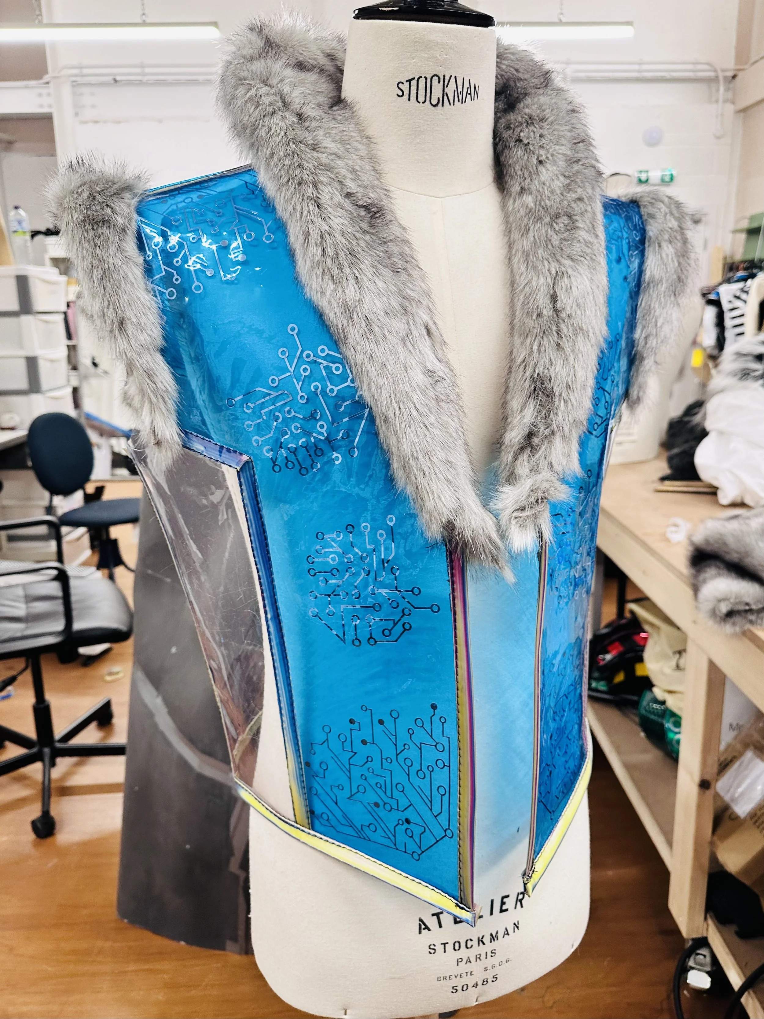 A fashion mannequin draped with a futuristic garment featuring a blue metallic panel with circuit-like patterns and gray fur trim on the shoulders and collar.