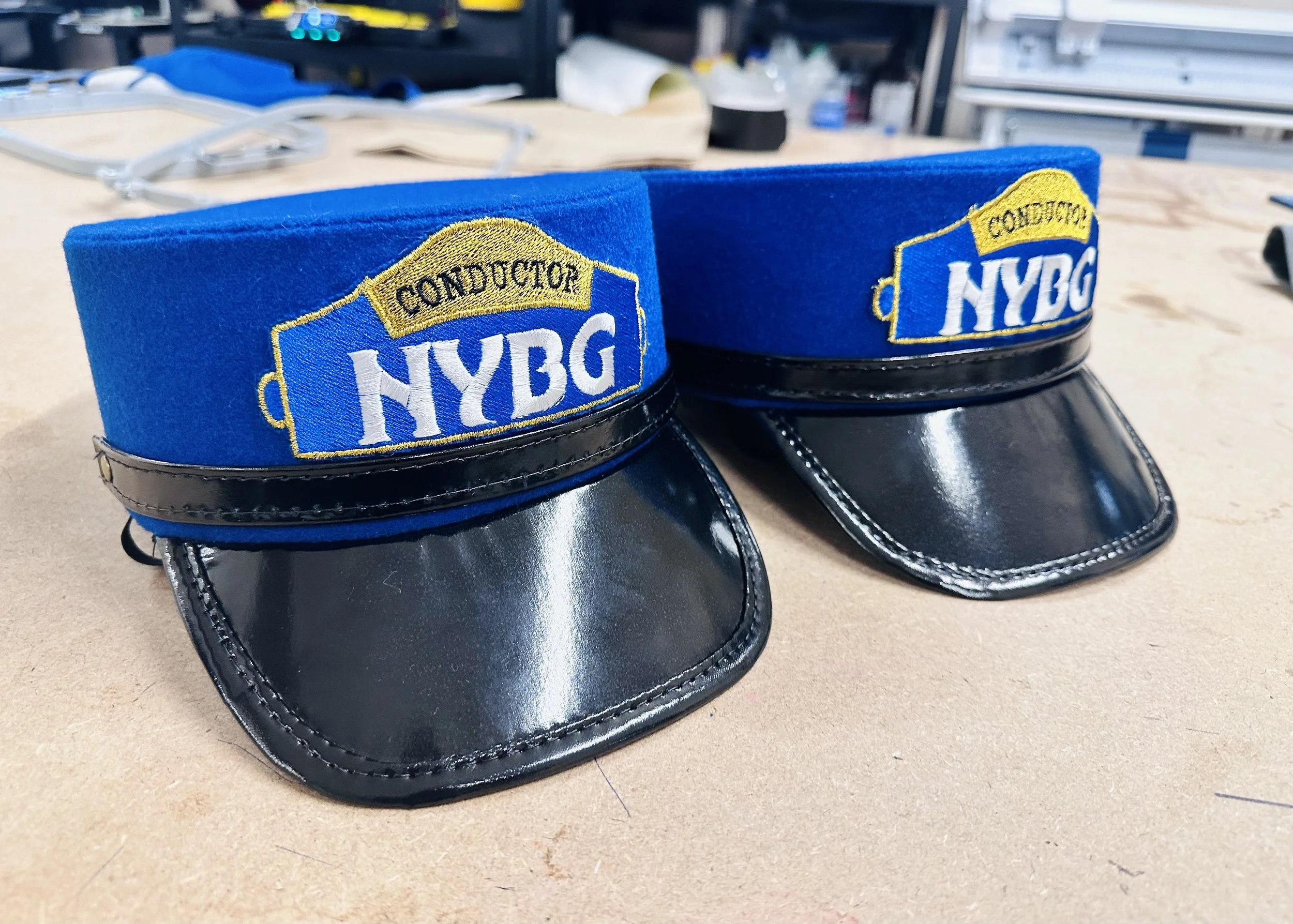 Two NYBDG conductor caps with blue fabric tops, yellow and white embroidered patches, and shiny black visors, placed on a workbench.