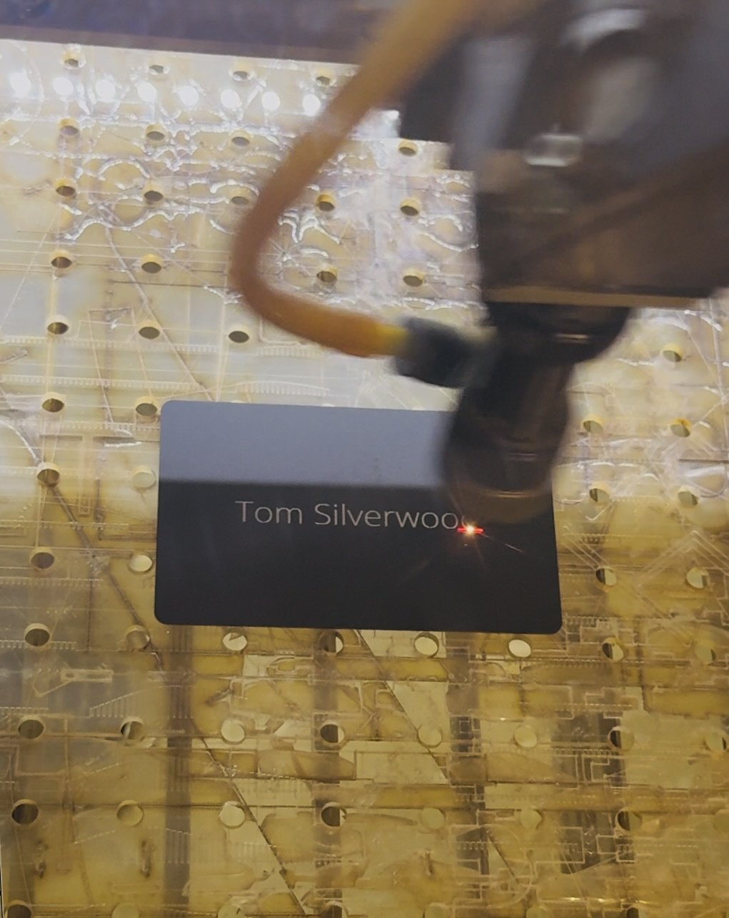 Close-up of a laser engraving machine working on a black rectangular object with the name 'Tom Silverwood' engraved on it, on a yellow perforated work surface.