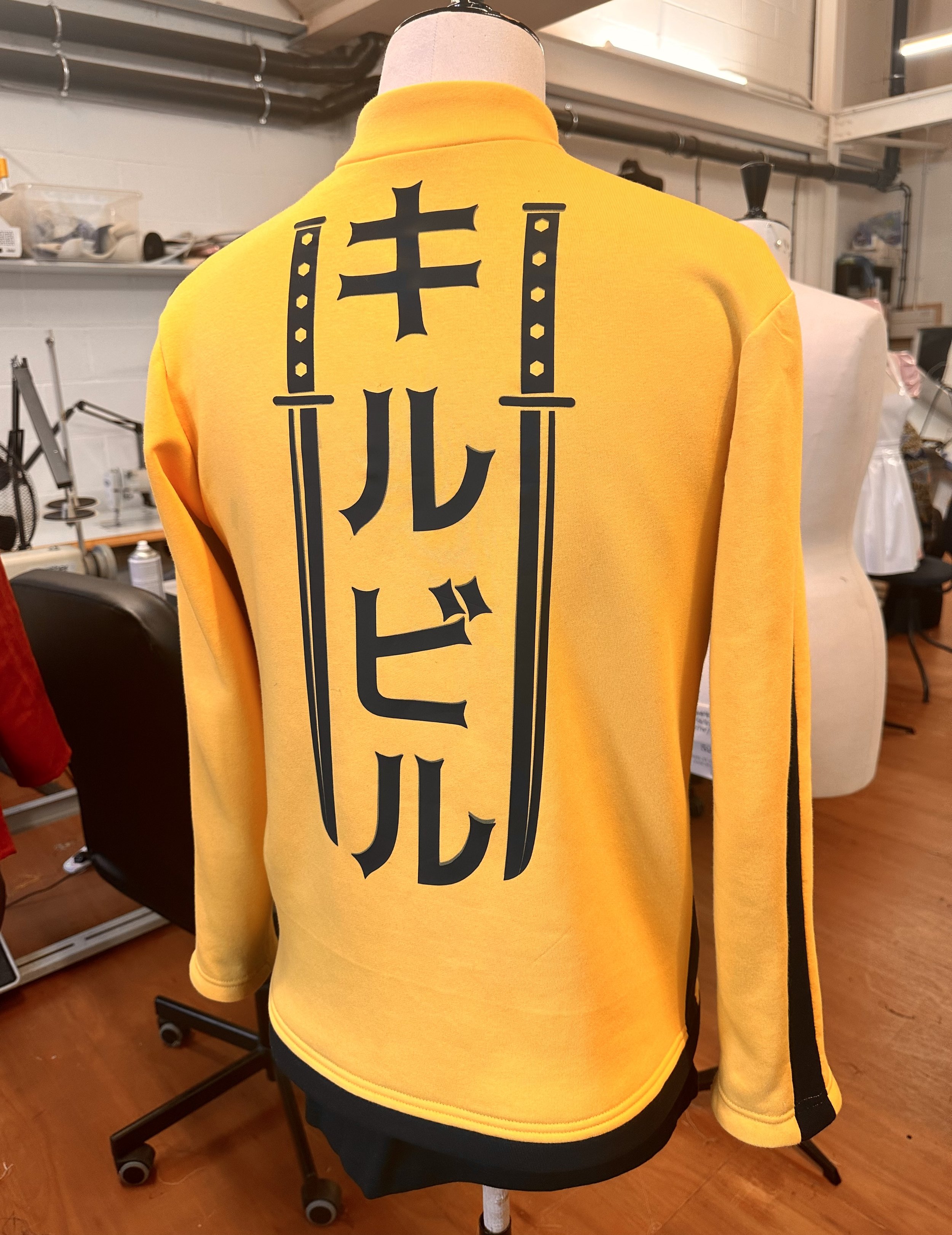 Yellow jacket with Japanese characters and sword graphics on the back, displayed on a dress form in a workshop.